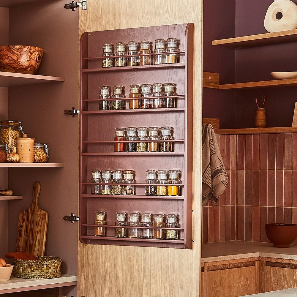 A wooden spice rack attached to the inside of a cabinet door holds several jars filled with herbs and spices in a modern kitchen with wood accents and pinkish tile backsplash.