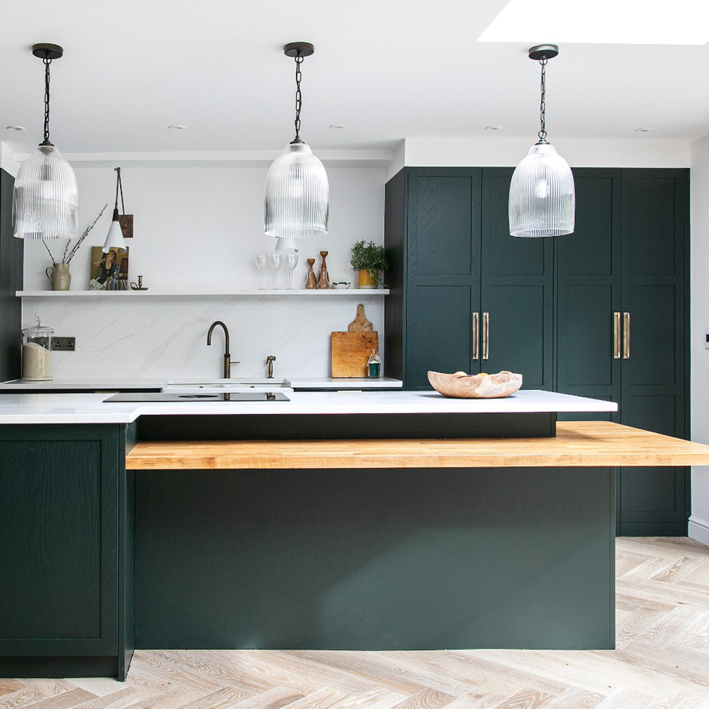 Modern kitchen with dark green cabinets, a white countertop, and a wooden breakfast bar extension. Three glass pendant lights hang above the island. Open shelves and decorative items are visible in the background.