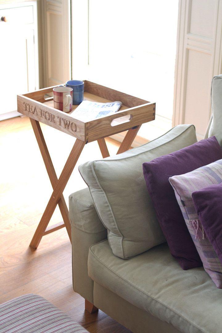 A wooden tray with 'tea for two' on it stands a folding stand next to light green couch with purple and striped pillows. The tray holds red and blue cups, newspaper. Room is softly lit by natural light.