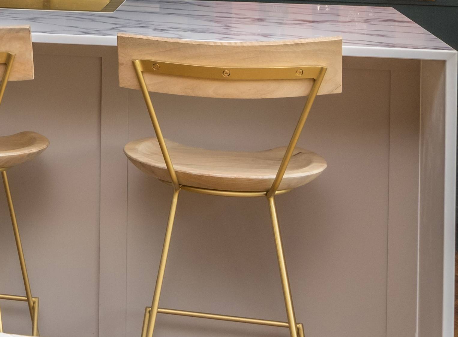 A modern wooden bar stool with a curved seat and backrest, supported by gold metal legs, positioned at a light-colored kitchen island with a marble countertop.