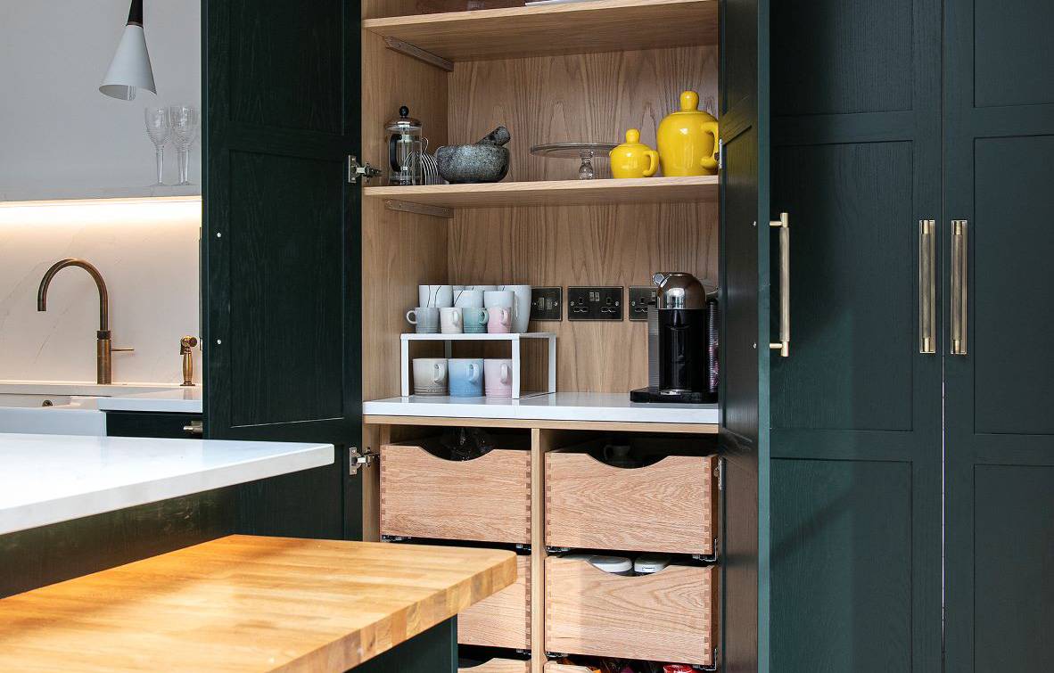 A modern kitchen with dark green cabinets and open pantry doors revealing shelves with mugs, a kettle, and a coffee maker. The countertop is white, and there's a wooden surface attached. The herringbone-patterned floor is light wood.