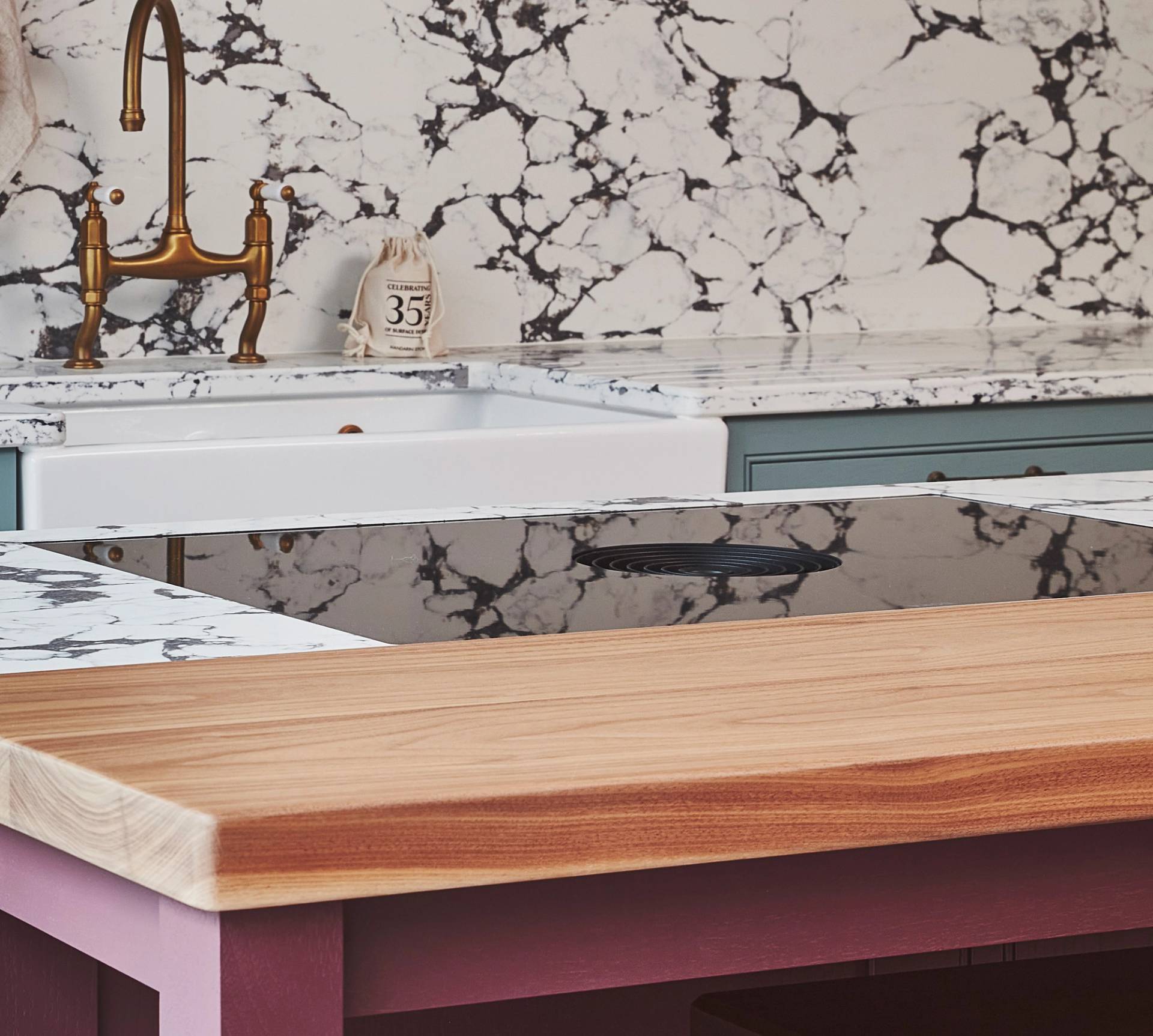 A kitchen counter area with a white marble backsplash featuring black veining, a farmhouse sink with a brass faucet, a blue cabinet, and a wooden countertop in the foreground. A decorative jar is near the sink