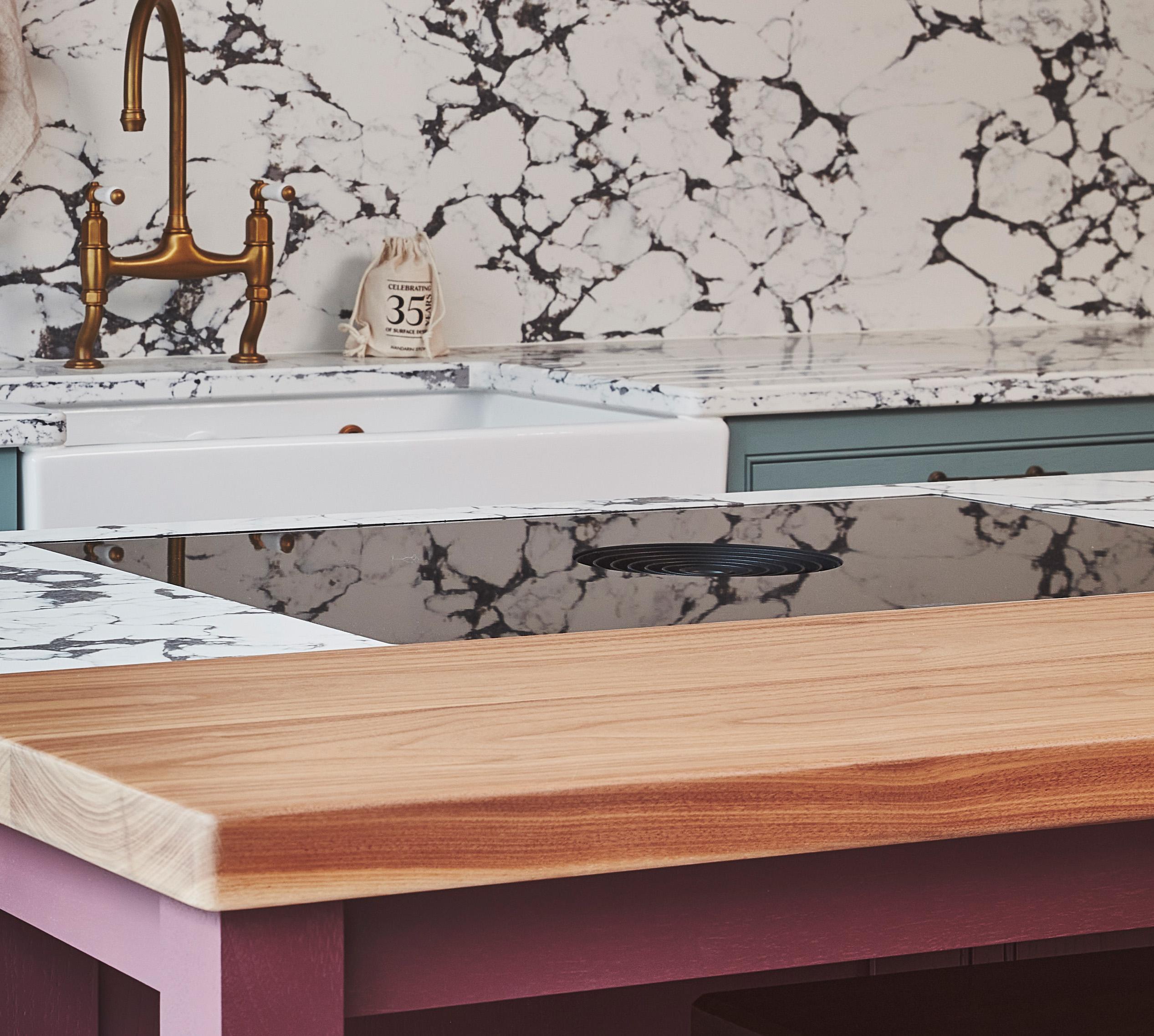 A kitchen counter area with a white marble backsplash featuring black veining, a farmhouse sink with a brass faucet, a blue cabinet, and a wooden countertop in the foreground. A decorative jar is near the sink