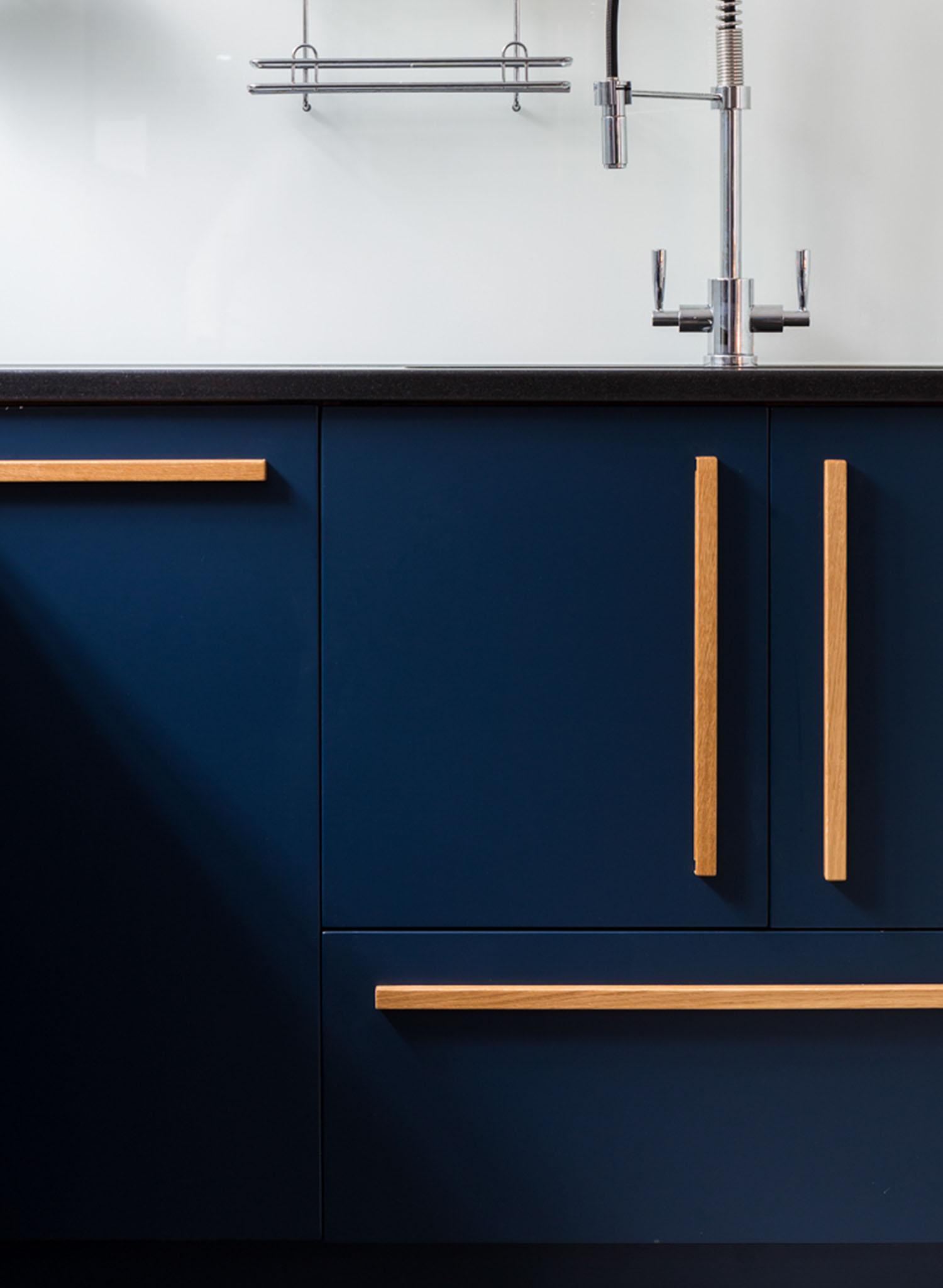 Modern kitchen cabinet with dark blue drawers and doors, featuring long, light wood handles. A silver faucet and a metal rack are visible above the black countertop, set against a white backsplash.