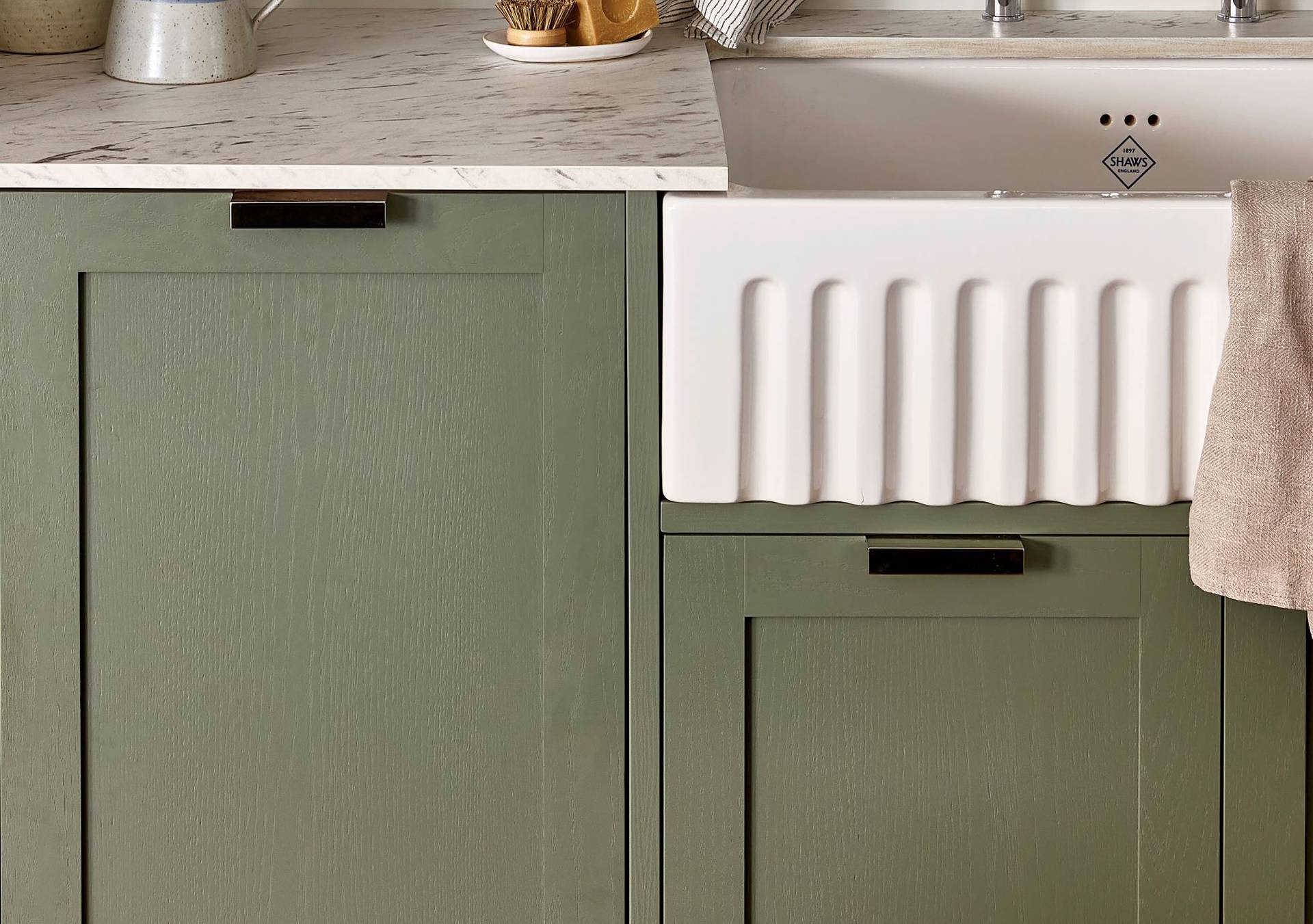 A kitchen setup features a marble countertop, pale green cabinets, and a white farmhouse sink with a ribbed front. A beige towel hangs over the sinks edge. A plant and a cleaning brush are on the counter.