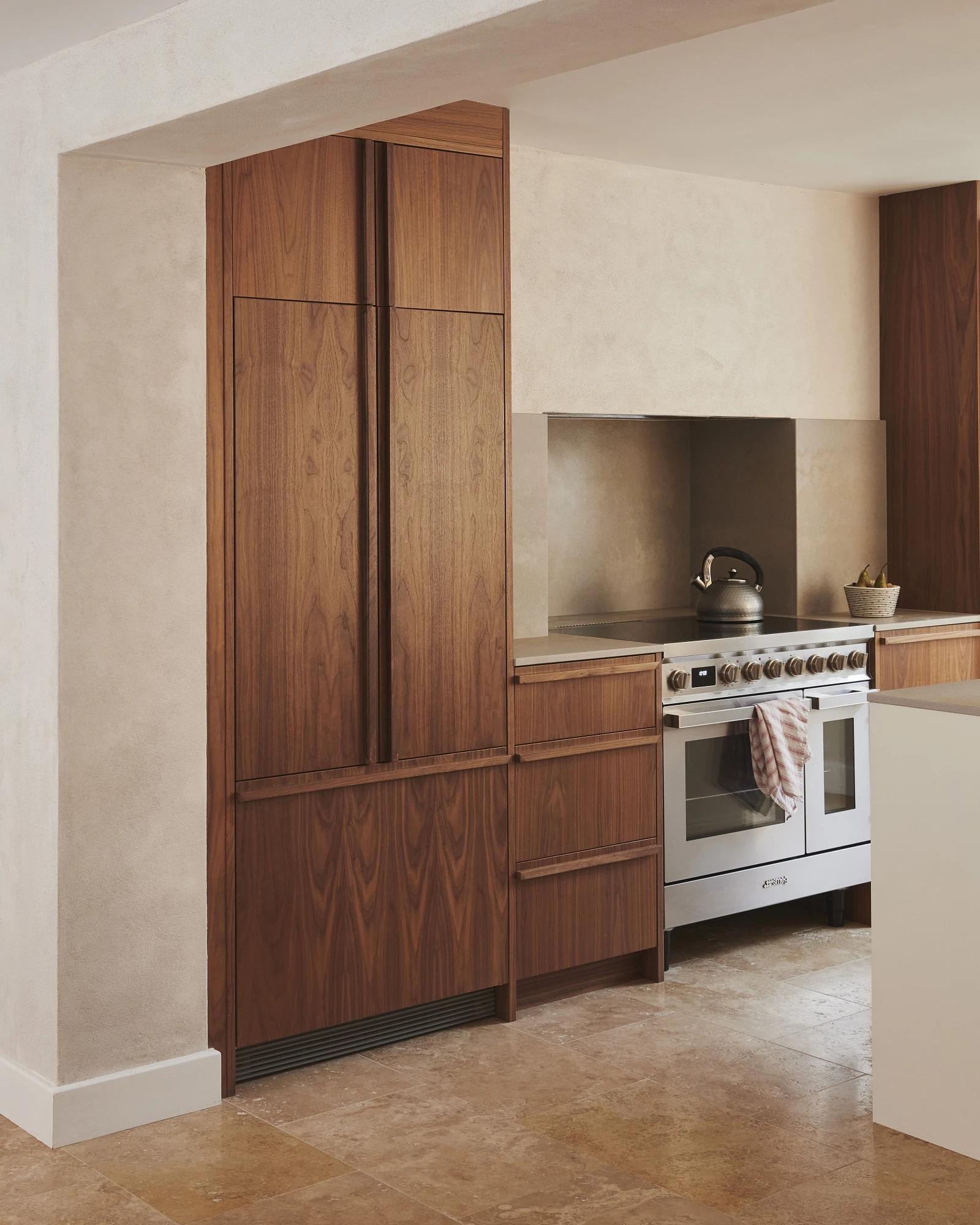 A modern kitchen with wooden cabinets, a stainless steel oven, and a white countertop. A kettle sits on the stovetop, and a small plant is placed on the counter next to the oven. The floor is made of light beige tiles.
