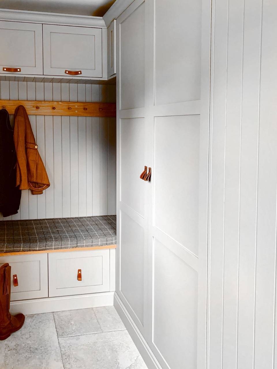 Mudroom with white paneled cabinets and a checkered bench. Hooks hold a brown jacket, and a pair of boots is placed nearby on the tiled floor. Brown leather handles accent the cabinets, adding a warm touch to the bright space.