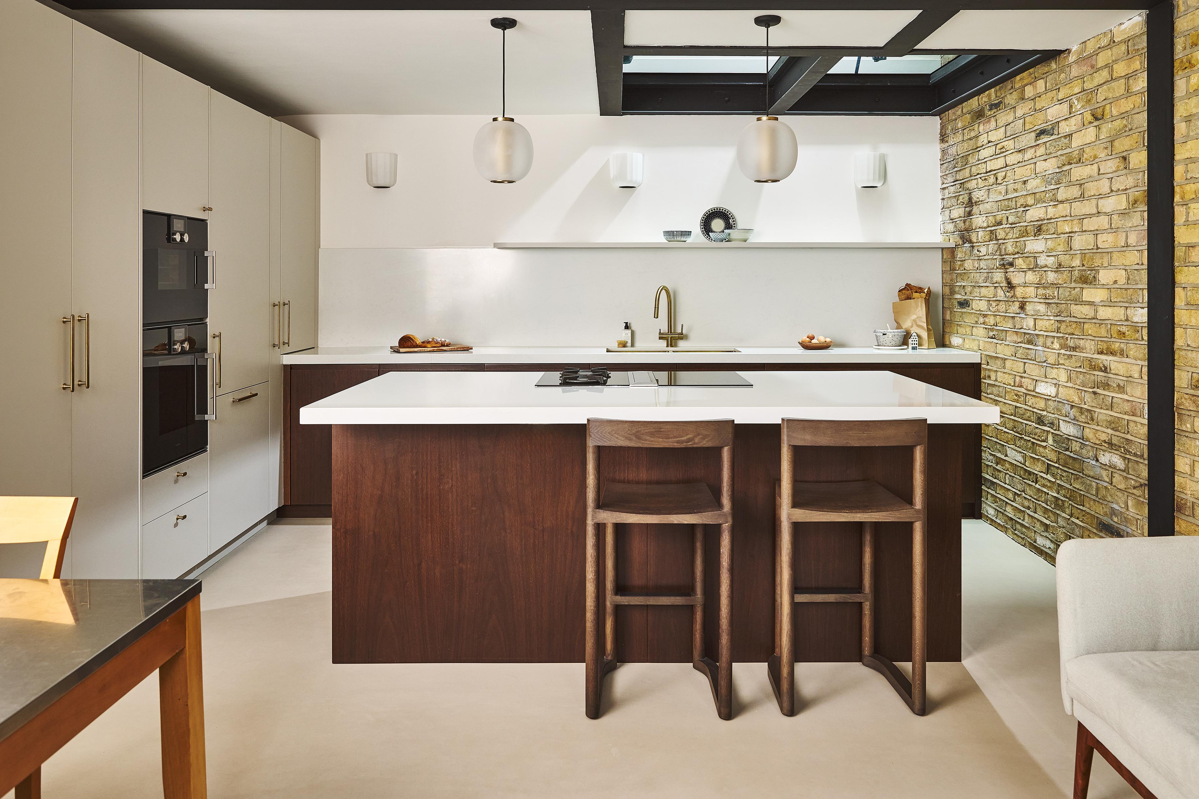 Modern kitchen with a central island, two wooden bar stools, white countertops, built-in ovens, minimalist cabinets, pendant lights, and an exposed brick wall on the right.