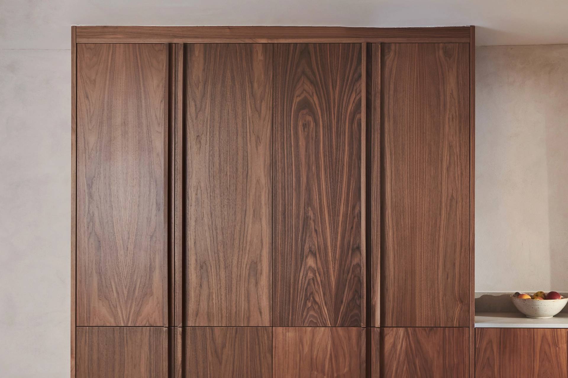 Wooden kitchen cabinets with a natural, rich grain pattern. A small section of a beige countertop is visible on the right, holding a white bowl filled with assorted fruit. The background wall is a light neutral color.
