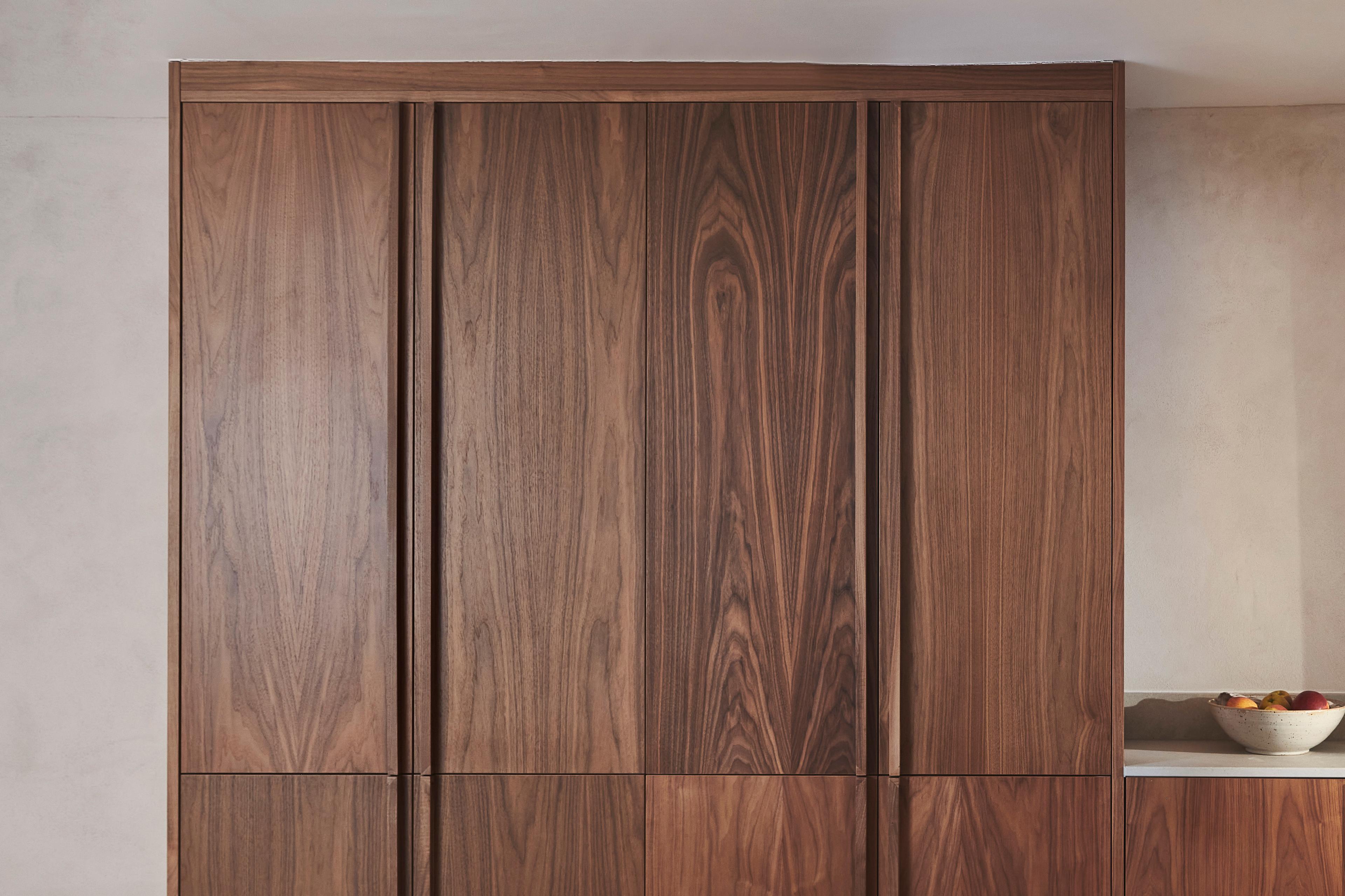 Wooden kitchen cabinets with a natural, rich grain pattern. A small section of a beige countertop is visible on the right, holding a white bowl filled with assorted fruit. The background wall is a light neutral color.