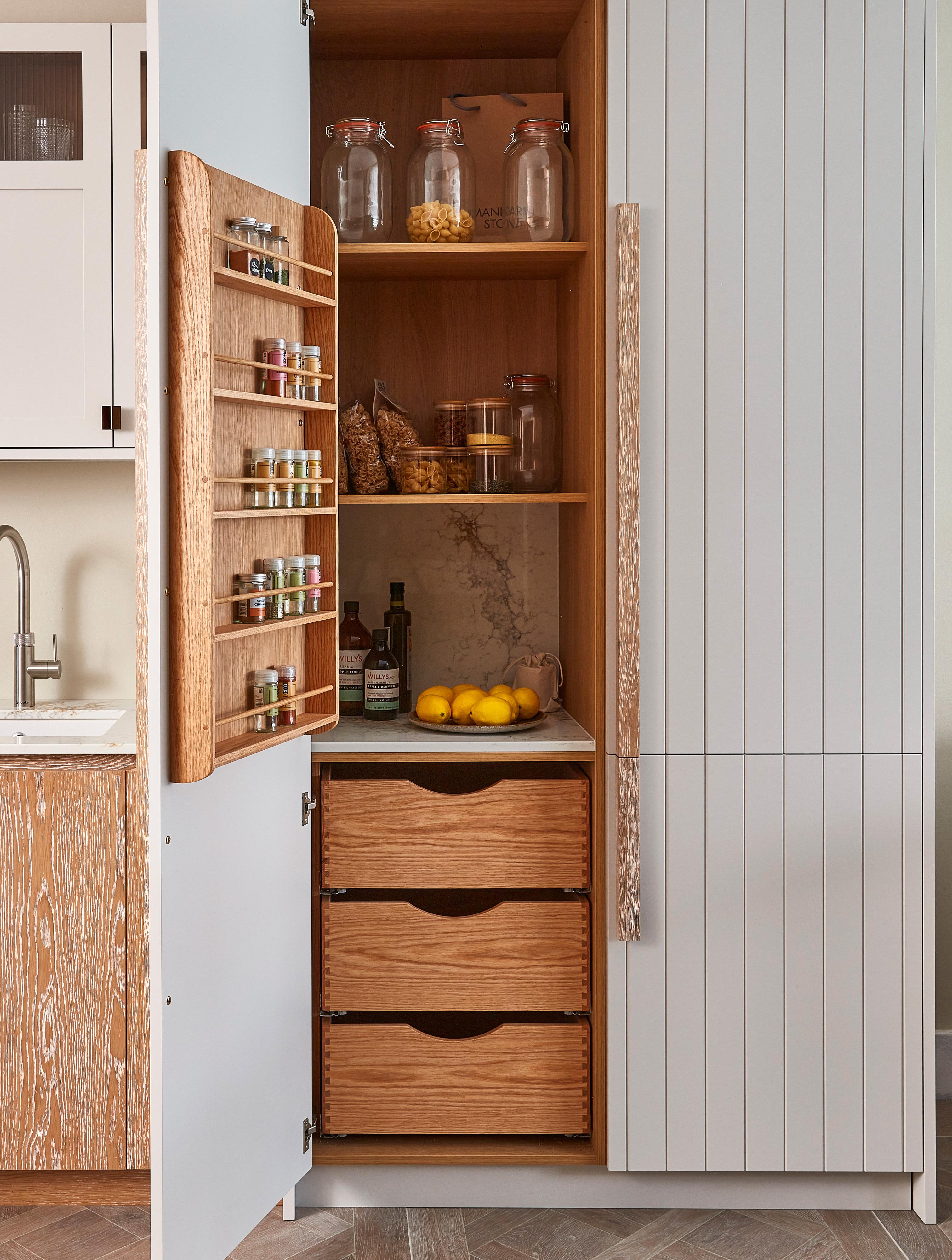 Open kitchen cabinet with a door-mounted spice rack, displaying small jars. Inside, glass jars with grains, a few lemons, and a bottle of olive oil are visible. Below are three large wooden drawers. Nearby is a kitchen sink.
