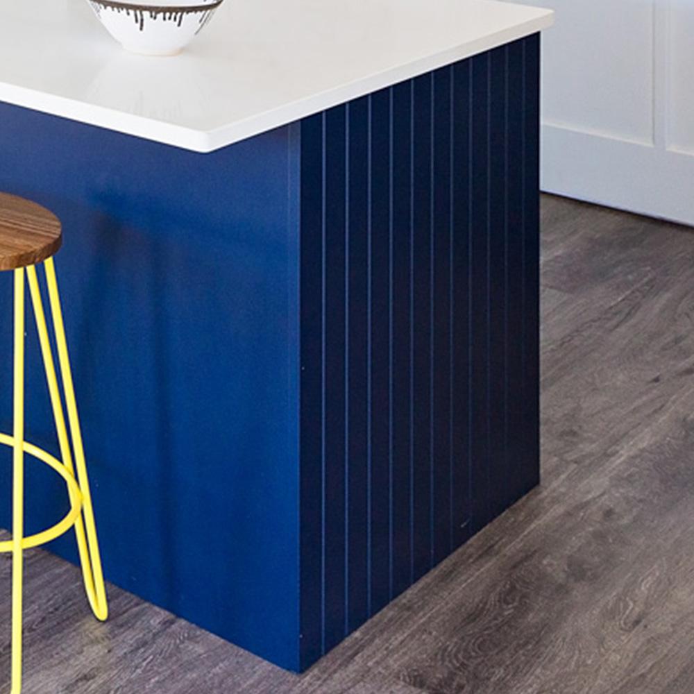 A modern kitchen island with a navy blue panel and white countertop, next to a wooden stool with bright yellow metal legs, set on gray wood-look flooring.