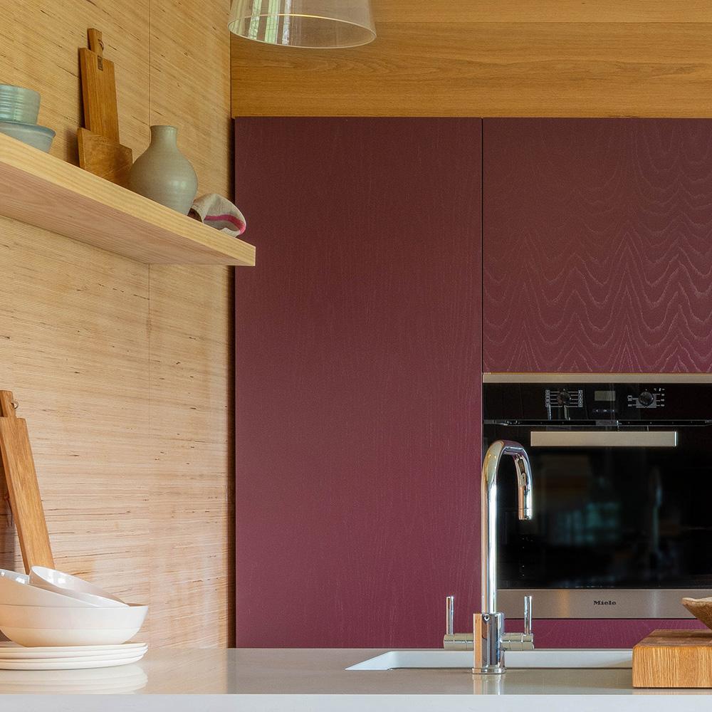 A modern kitchen with maroon cabinets, a built-in oven, a sleek faucet, light wood walls, and a floating shelf holding bowls, plates, vases, and cutting boards.