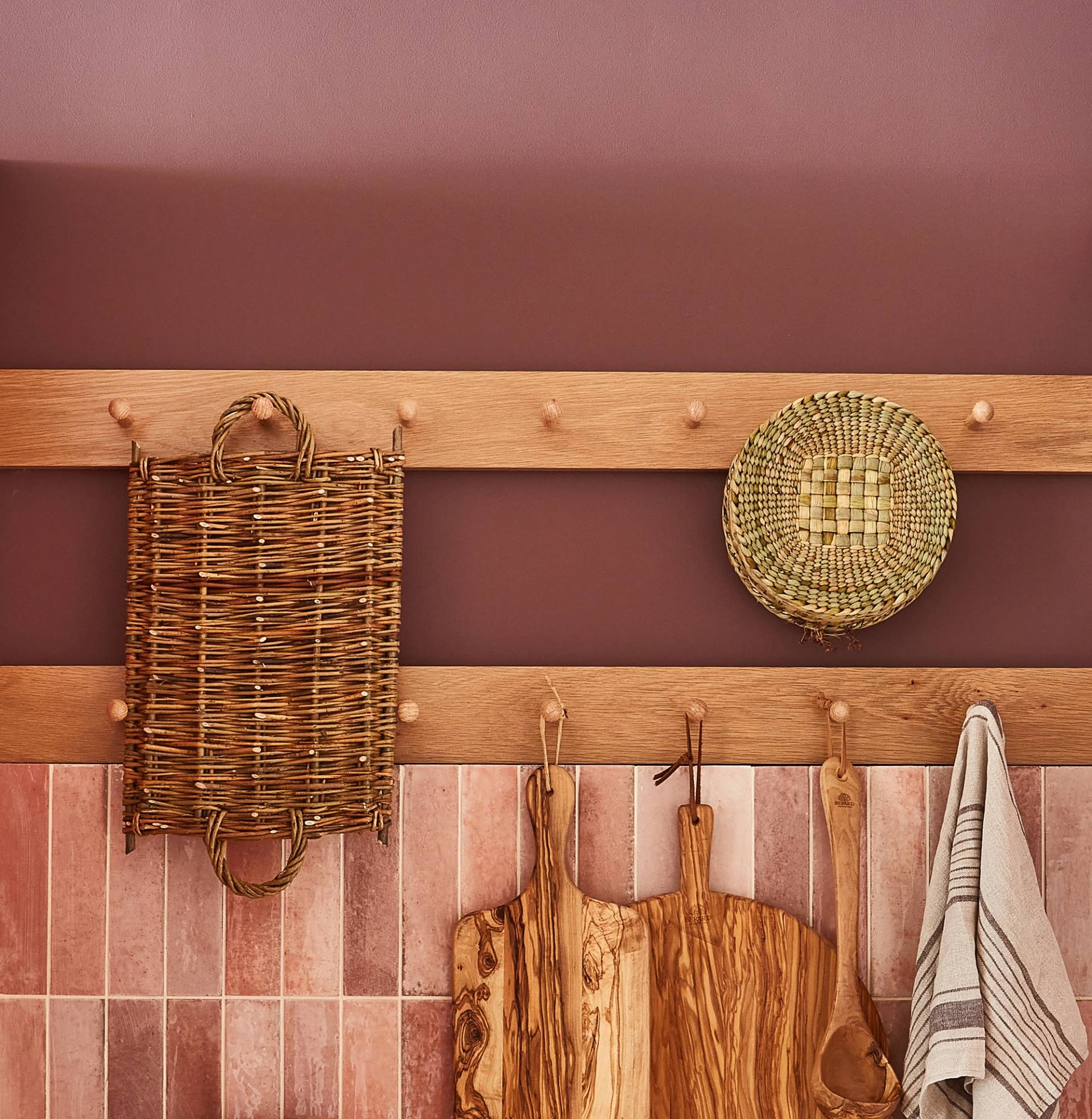 A wooden wall with pegs holds woven baskets, wooden cutting boards, and a striped kitchen towel against a warm-toned background. The tile and wood textures add a rustic charm