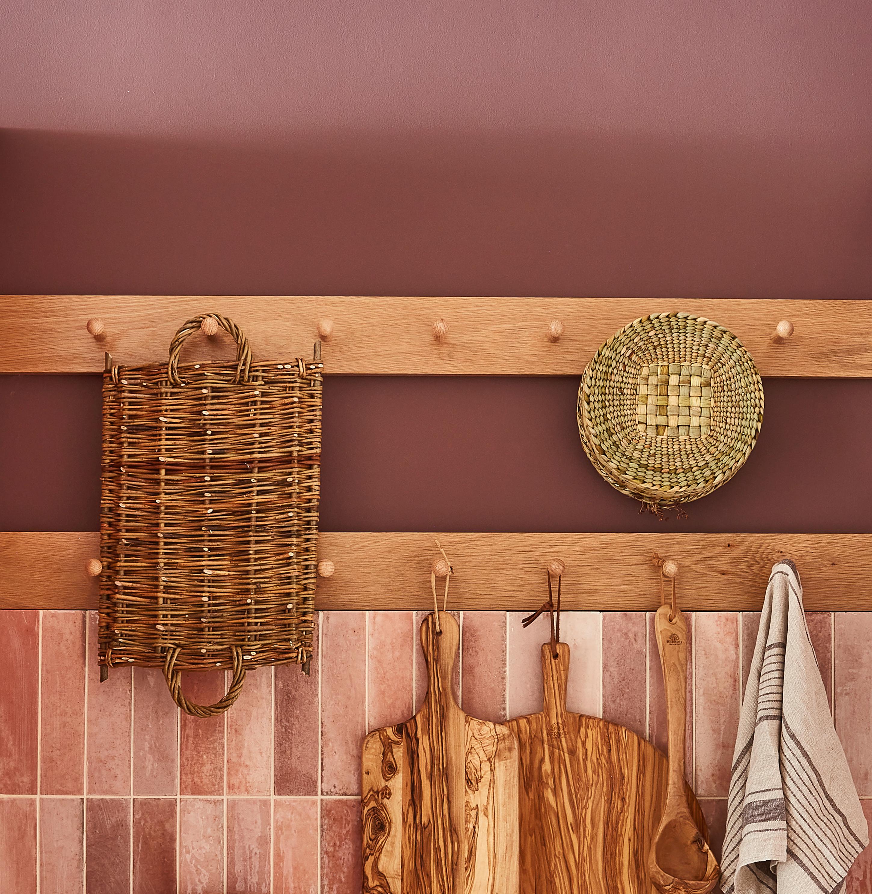A wooden wall with pegs holds woven baskets, wooden cutting boards, and a striped kitchen towel against a warm-toned background. The tile and wood textures add a rustic charm