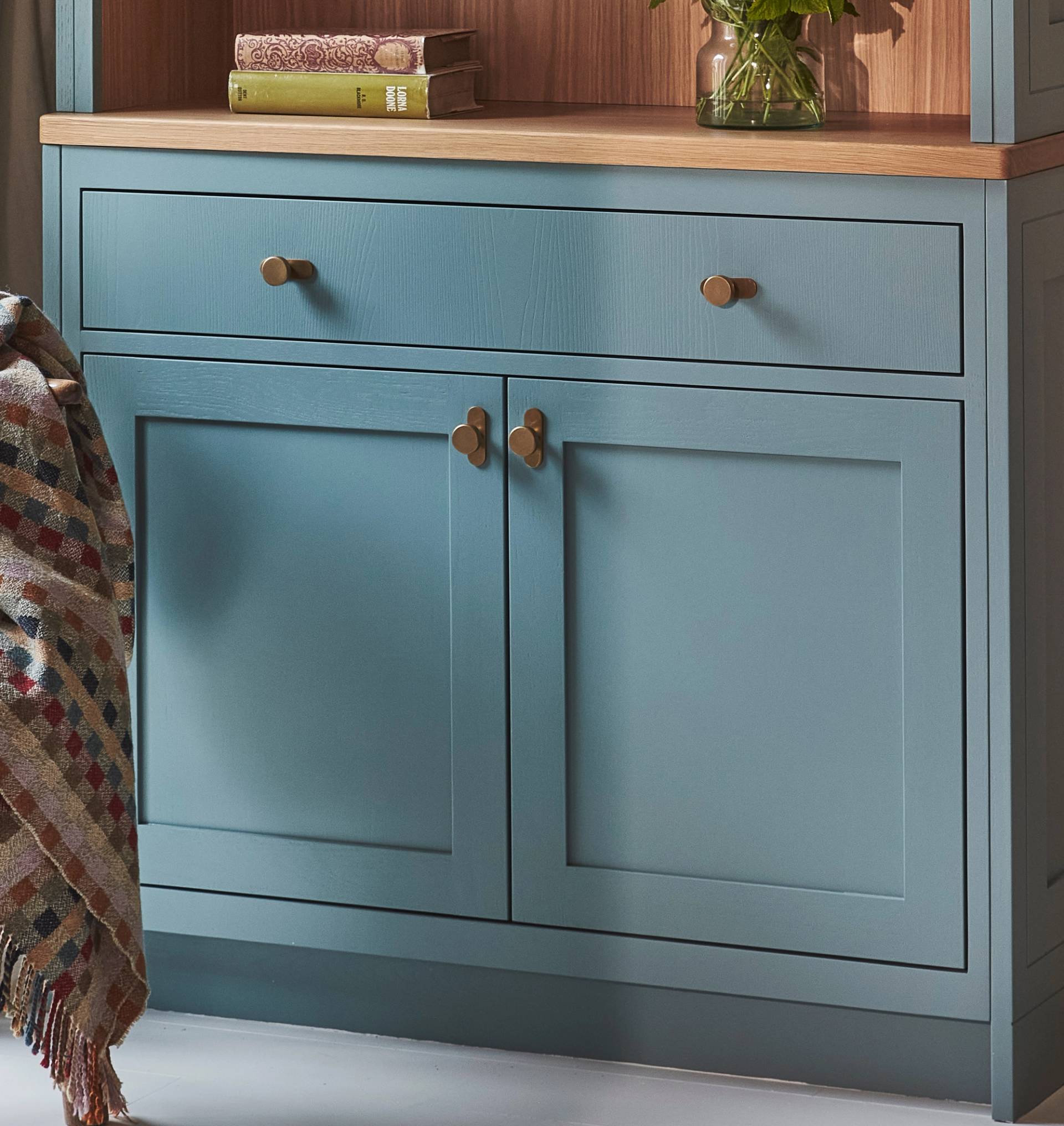 A blue wooden cabinet with two drawers and two doors, featuring brass knobs. On top, there are books and a vase with flowers. A multicolored patterned blanket is draped over the left side.