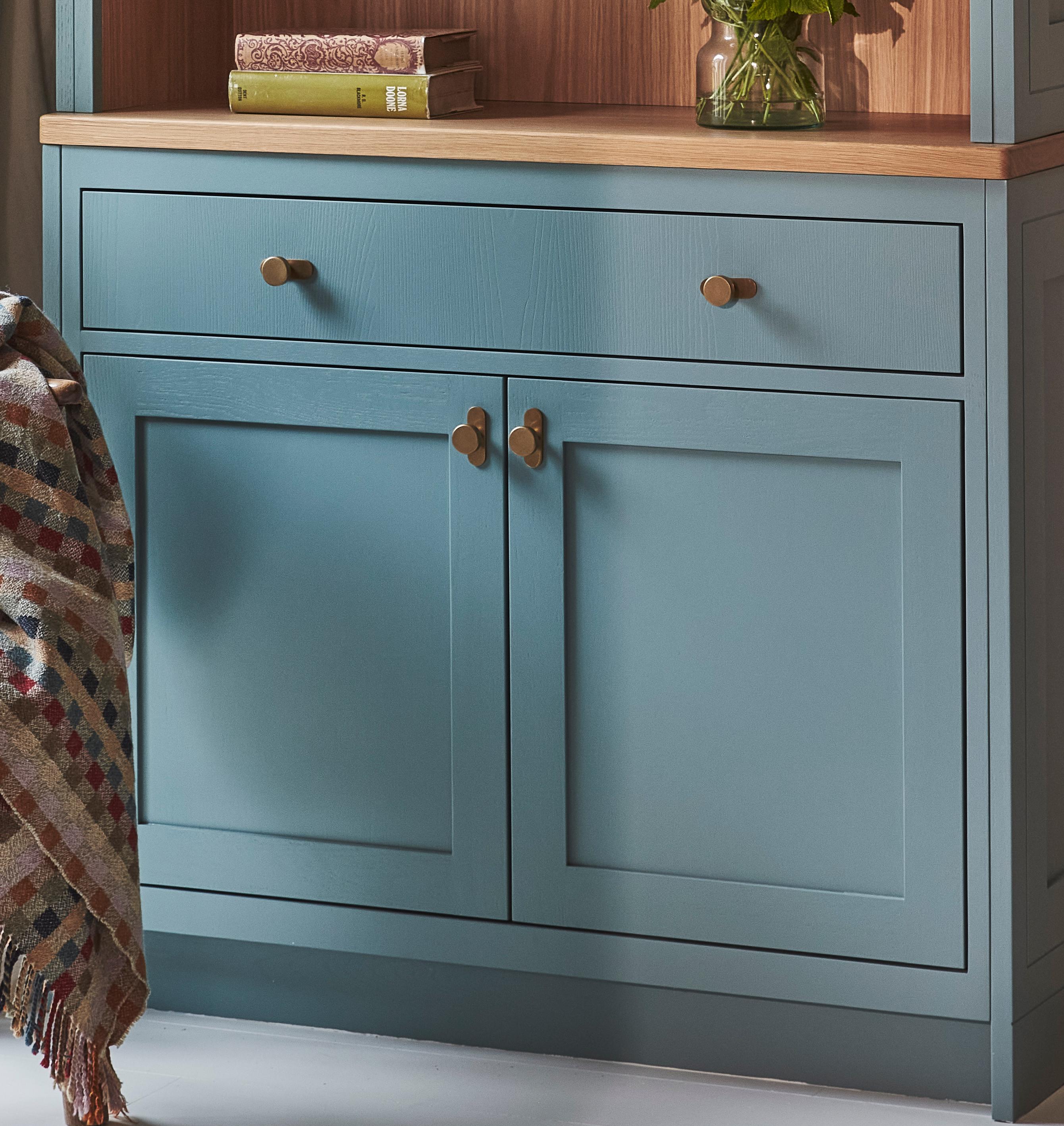 A blue wooden cabinet with two drawers and two doors, featuring brass knobs. On top, there are books and a vase with flowers. A multicolored patterned blanket is draped over the left side.