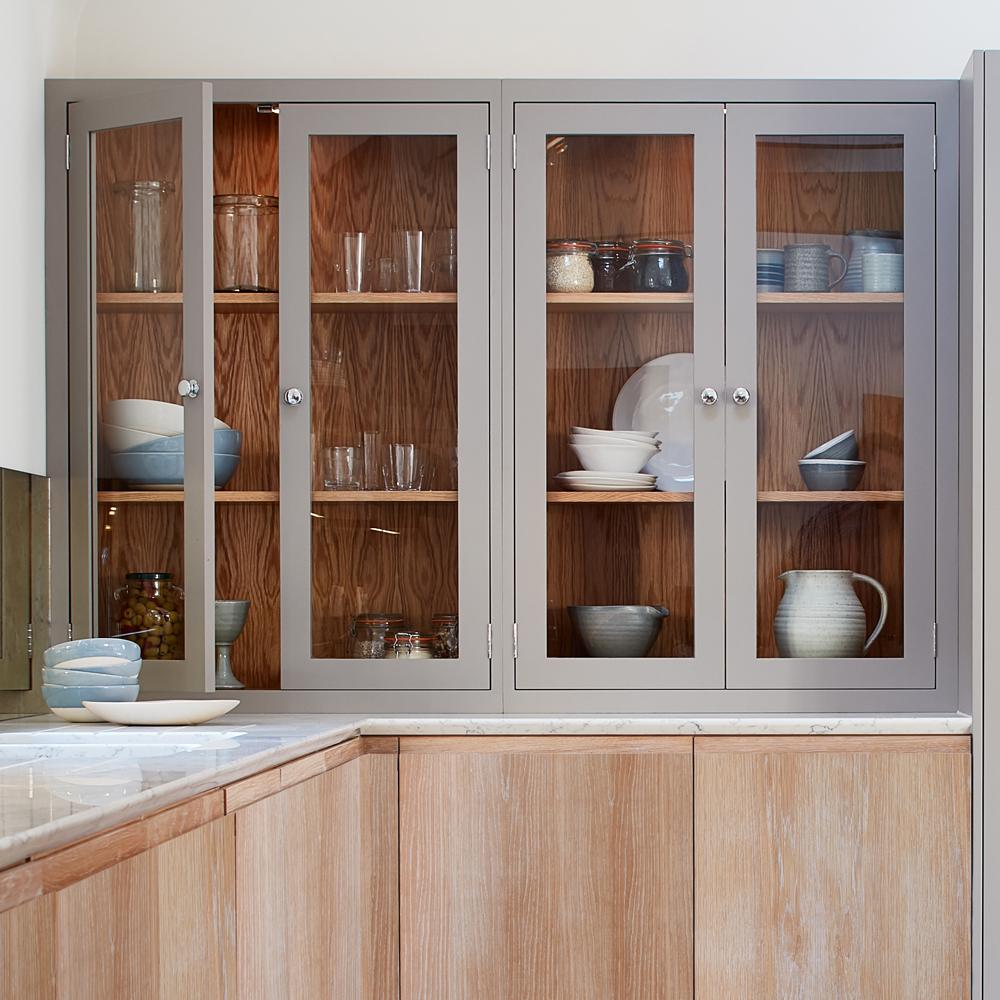 A set of glass-fronted kitchen cabinets displays neatly arranged dishes, bowls, glasses, and pitchers on wooden shelves. One cabinet door is slightly open, revealing the organized contents inside.