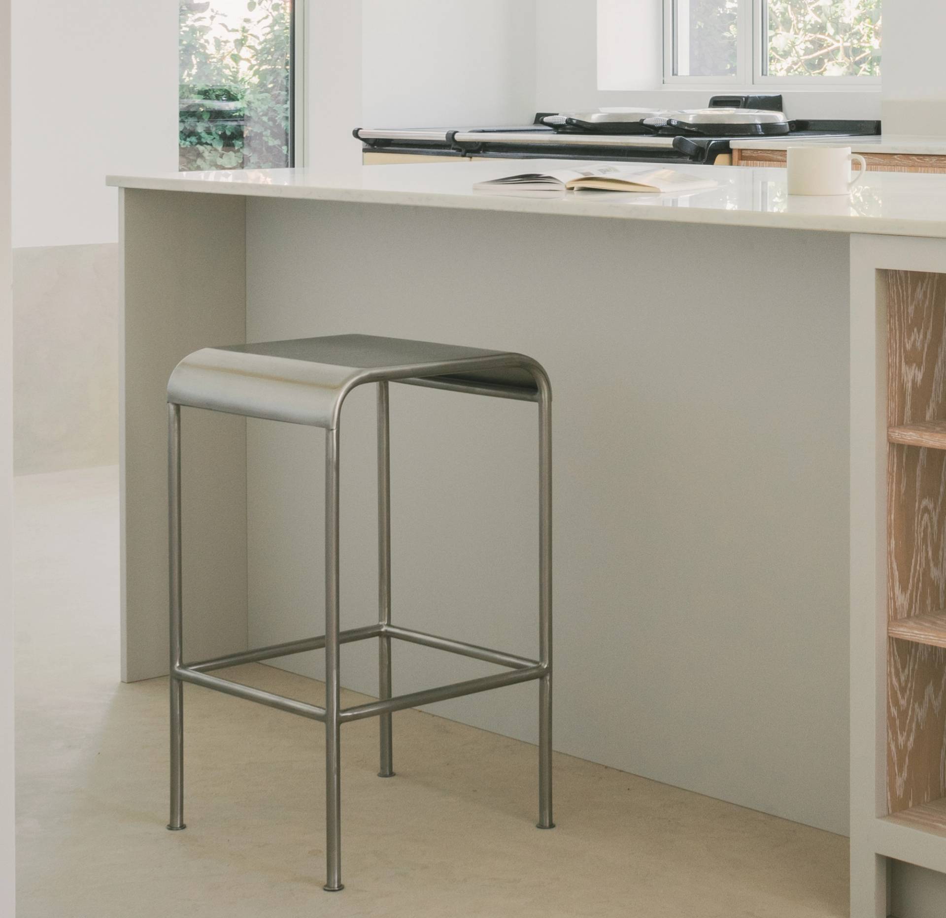 A modern kitchen with a minimalist metal stool placed at a white island countertop. An open book and a cup sit on the counter, with natural light coming through a window in the background.