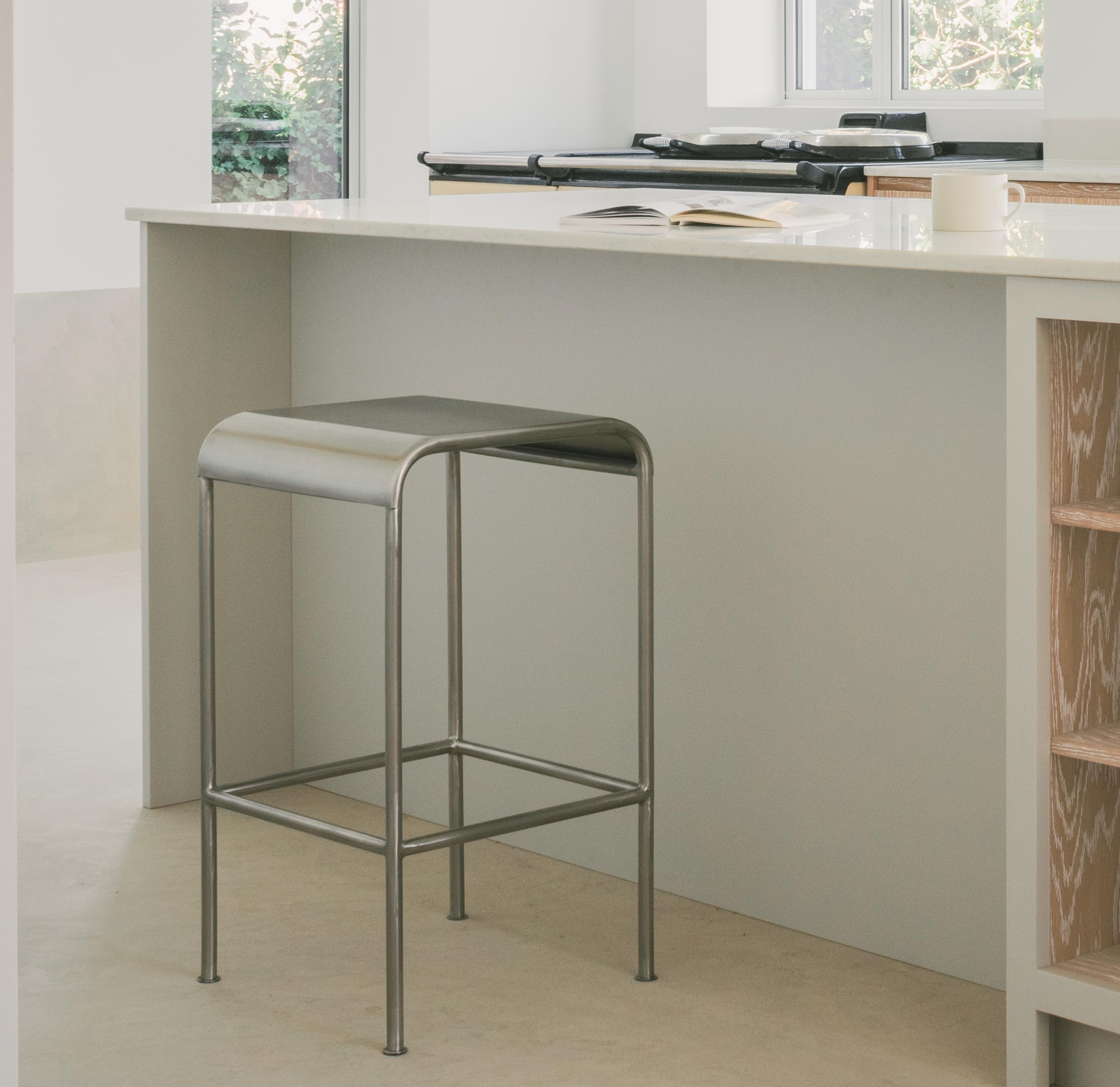A modern kitchen with a minimalist metal stool placed at a white island countertop. An open book and a cup sit on the counter, with natural light coming through a window in the background.