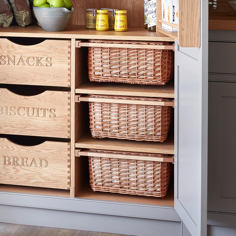 A kitchen storage unit with labeled wooden drawers for snacks, biscuits, and bread, alongside shelves holding woven wicker baskets and jars on the countertop above.
