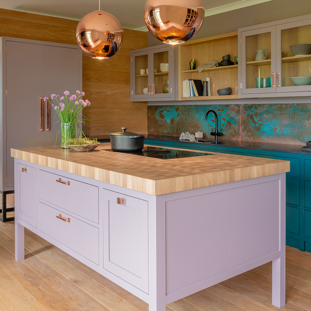 A modern kitchen with pastel purple cabinets, a wooden countertop island, copper pendant lights, and teal lower cabinets. Glass-front upper cabinets display dishes, and a vase of flowers sits on the island