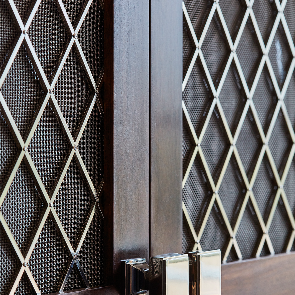Close-up of a wooden door with metal mesh panels arranged in a diamond pattern. The door has shiny silver handles and the background shows a dark, textured screen behind the mesh.