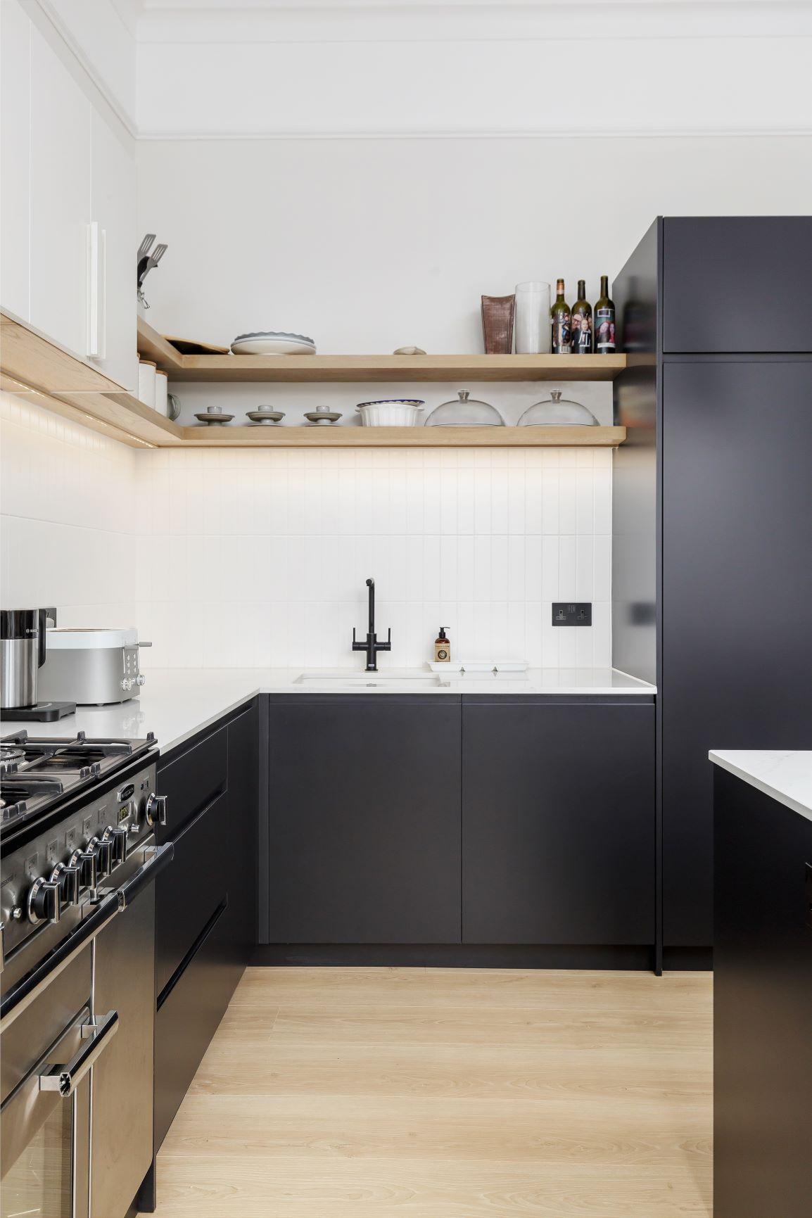 Modern kitchen with black lower cabinets, white countertops, and light wood shelves holding dishes and bottles. There’s a gas stove, a black faucet, and light wood flooring. The overall design is minimalist and tidy.