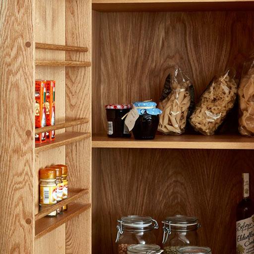 Wooden pantry shelves holding jars of spices, pasta in plastic bags, jam jars, and glass jars filled with dry goods. The shelves are neatly organized against a wood-grain background.