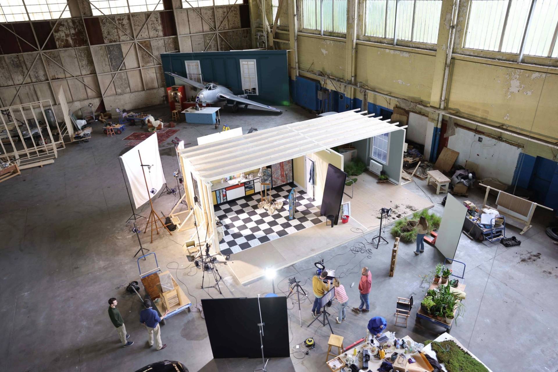 An expansive, high-angle wide shot of a large film studio or warehouse containing multiple active production sets. In the center, a crew works around a modular set featuring a black-and-white checkered floor and Mondrian-style cabinetry. Lighting rigs, cameras, and monitors surround the structure. In the background, another set features a silver vintage fighter jet integrated into a teal wall. The industrial space is filled with equipment, props, and various team members collaborating on the different stages of the shoot.