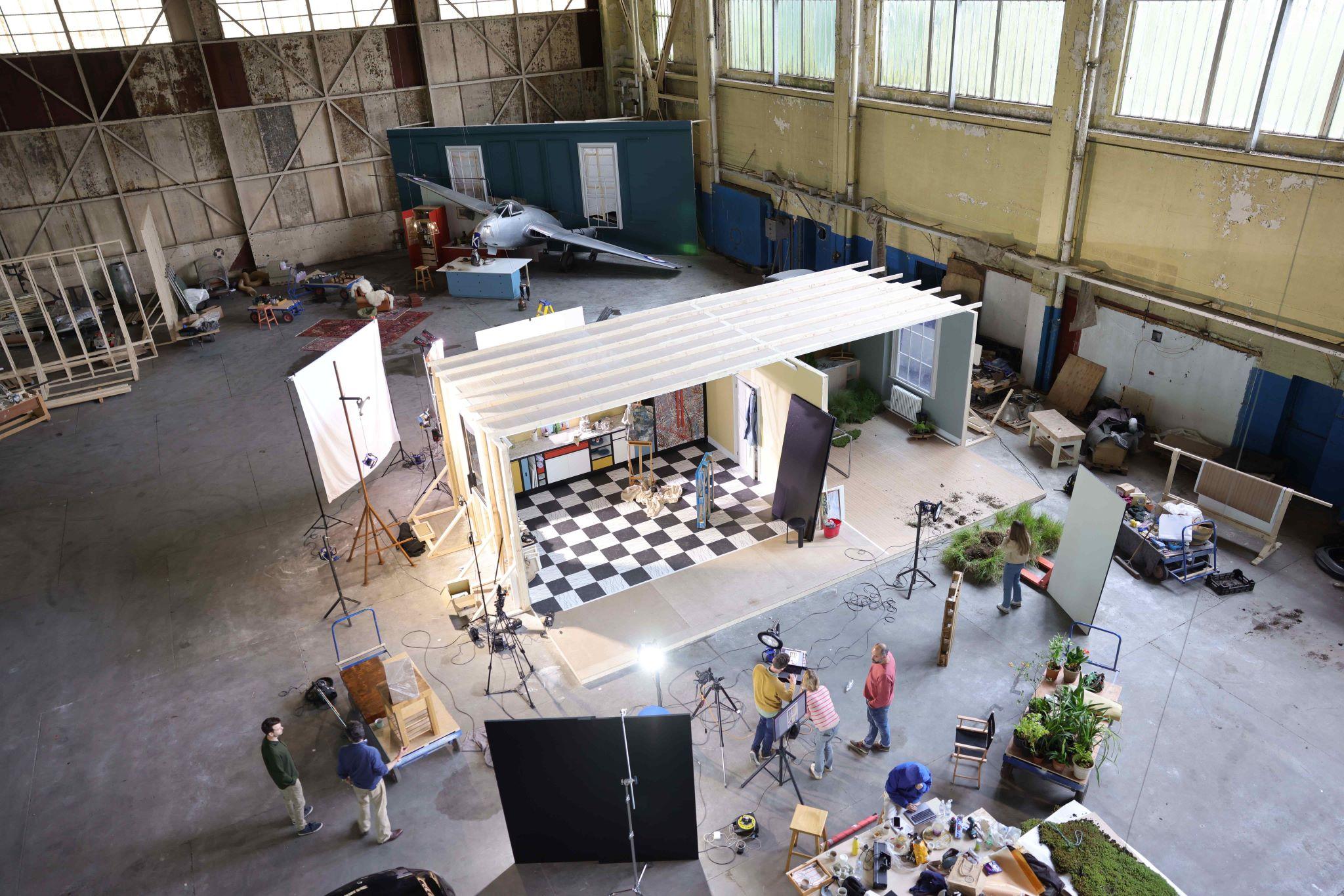 An expansive, high-angle wide shot of a large film studio or warehouse containing multiple active production sets. In the center, a crew works around a modular set featuring a black-and-white checkered floor and Mondrian-style cabinetry. Lighting rigs, cameras, and monitors surround the structure. In the background, another set features a silver vintage fighter jet integrated into a teal wall. The industrial space is filled with equipment, props, and various team members collaborating on the different stages of the shoot.