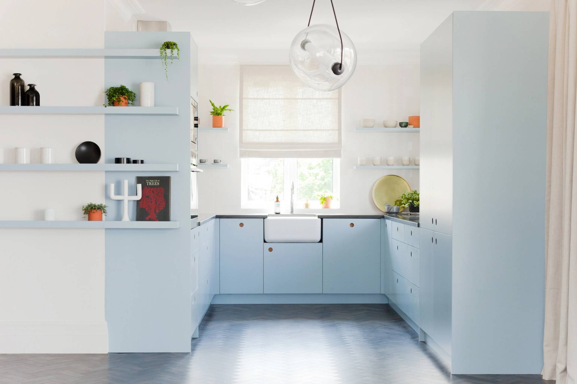 Bright, modern kitchen with pale blue cabinets, a farmhouse sink, white walls, open shelves holding dishes and plants, a window with a sheer white blind, and a dark herringbone-patterned floor.