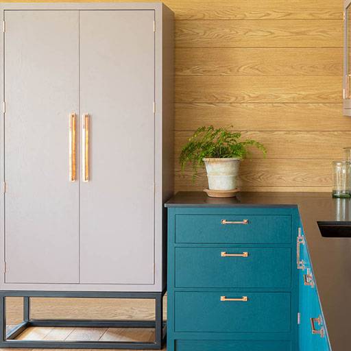 A modern room corner featuring a gray wardrobe with wooden handles on the left and a teal chest of drawers on the right. A fern in a pot is placed atop the chest, against a backdrop of light wood paneling.