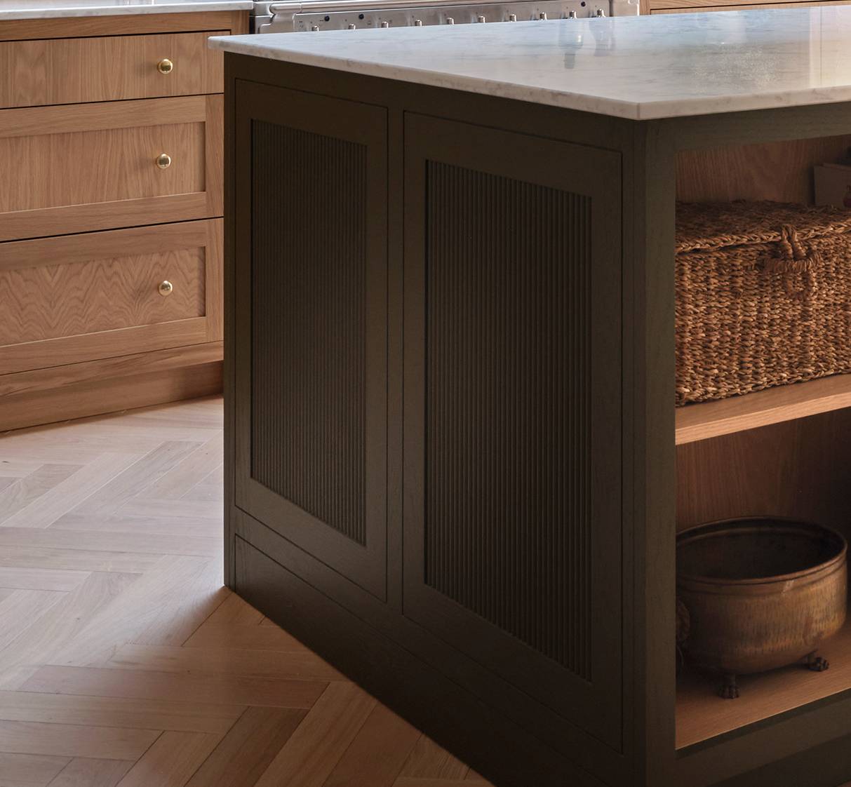 A kitchen island with dark paneling and a white countertop, open shelving with a woven basket and a metal bowl, and light wood cabinets with brass knobs in the background.