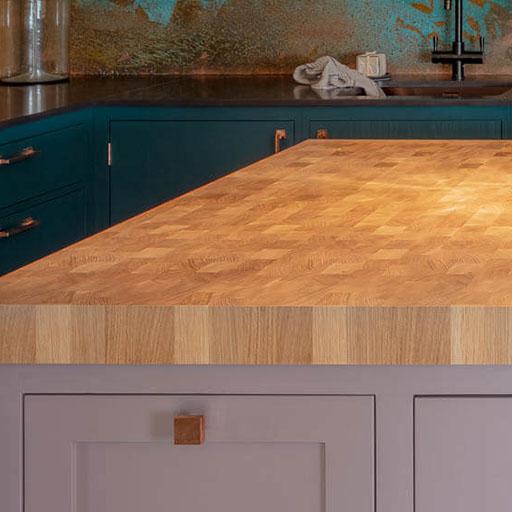 A kitchen island with a light wooden butcher block countertop, light gray cabinets, and dark teal cabinets in the background. A towel is draped near a black sink and faucet.