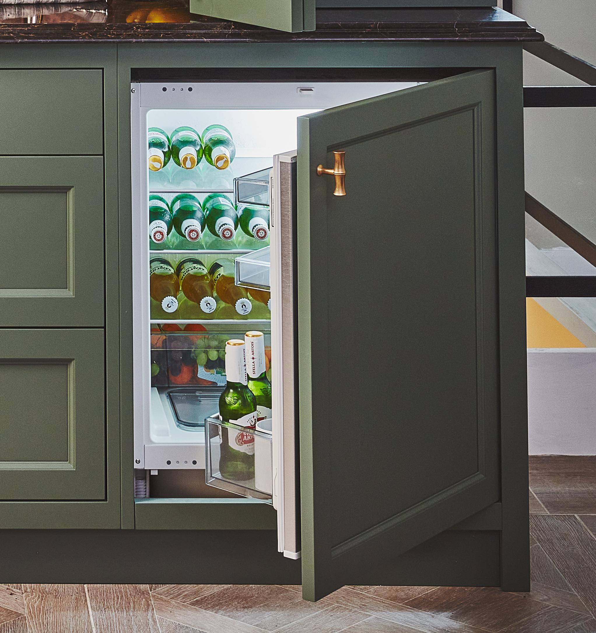 A built-in under-counter fridge with the door open, revealing shelves stocked with green glass bottles and a few items in the door compartment. The cabinetry is dark green with a brass handle.
