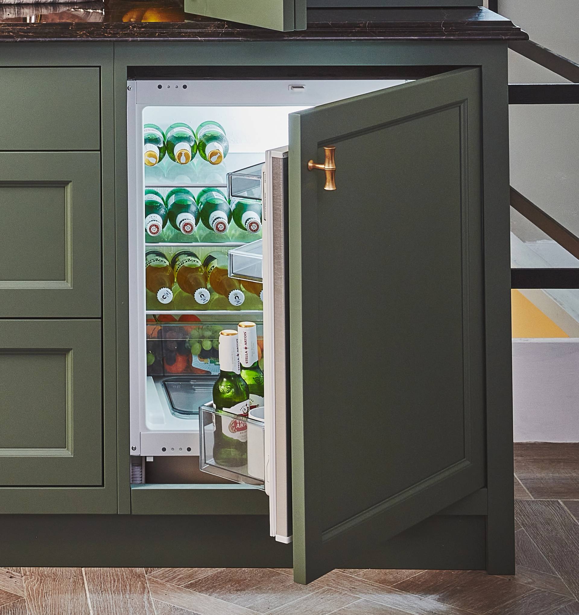 A built-in under-counter fridge with the door open, revealing shelves stocked with green glass bottles and a few items in the door compartment. The cabinetry is dark green with a brass handle.
