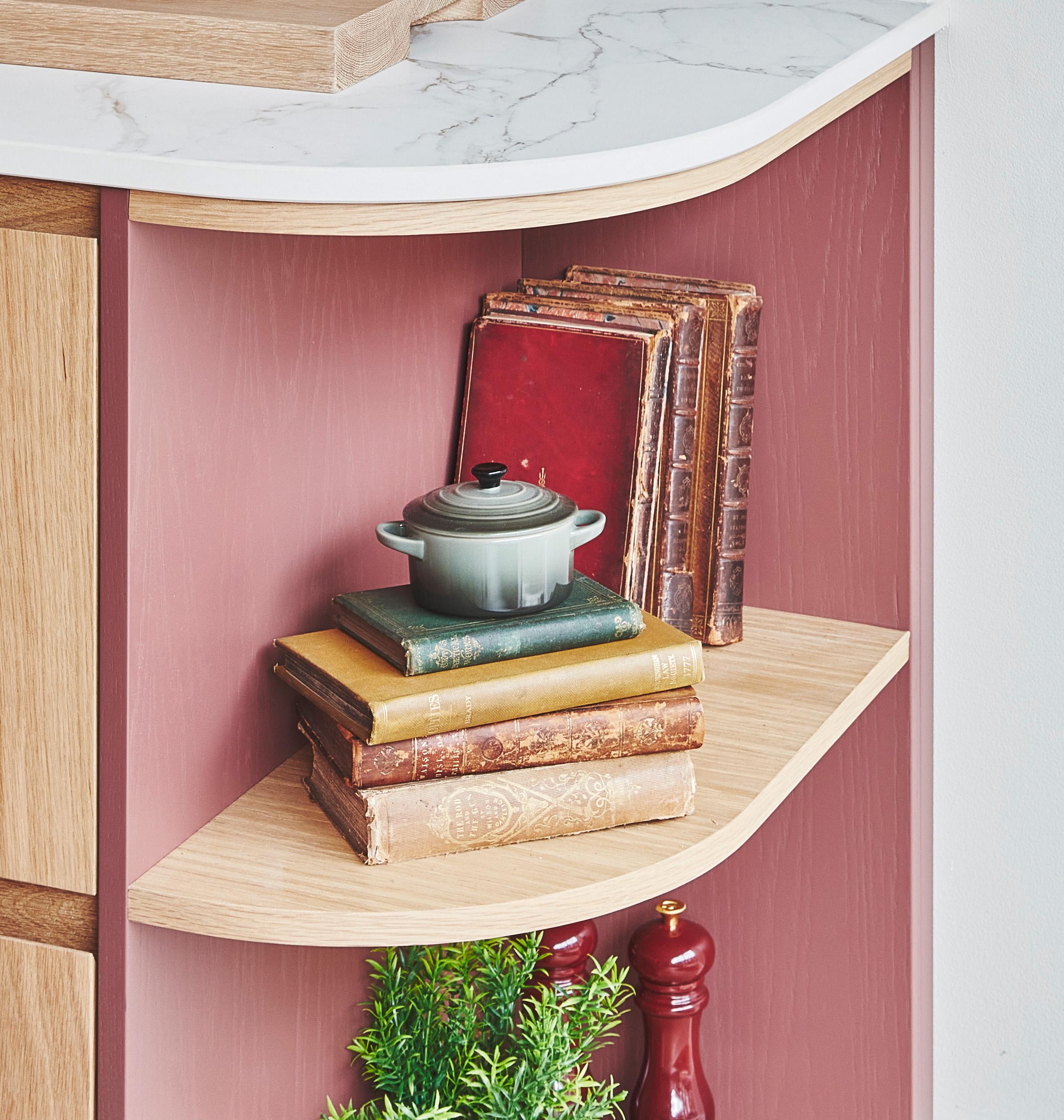 A small shelf with a stack of old books, a miniature gray pot on top, and red pepper grinders below. A small green plant adds a touch of nature to the cozy corner. The shelf has a marble-effect top.
