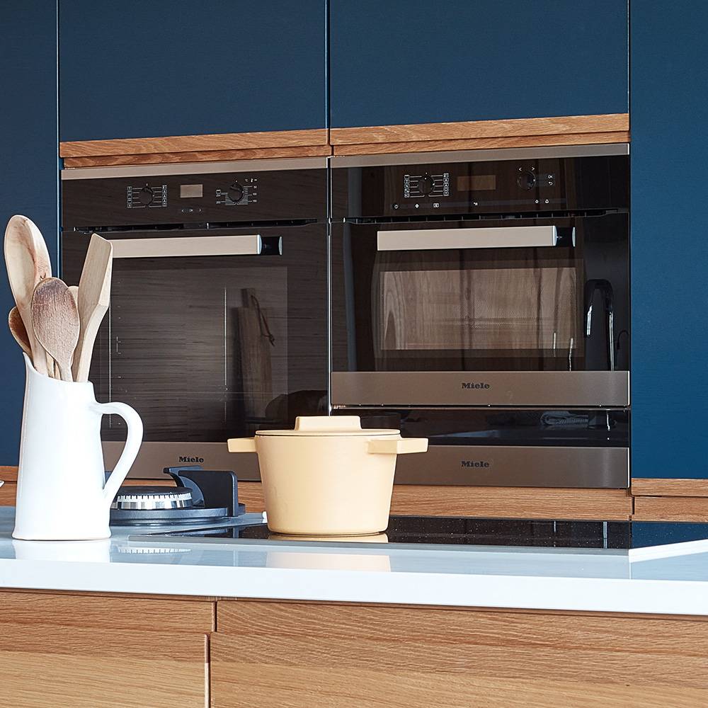 A modern kitchen with blue cabinets, built-in black and stainless steel ovens, a beige pot on the counter, and a white jug holding wooden utensils.