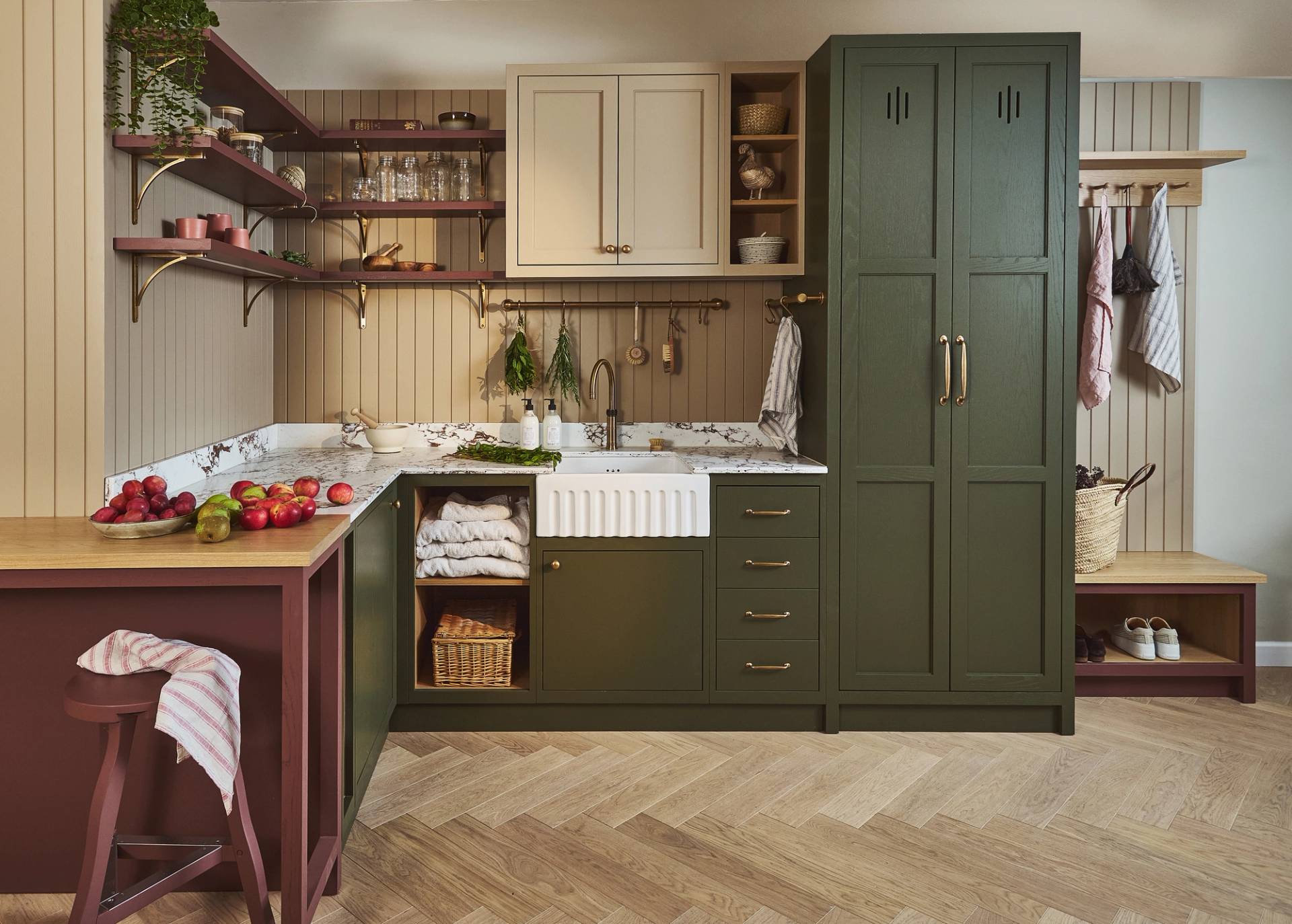 utility room with dark green and maroon cabinetry. There is a sink and tap in centre and fresh herbs hanging from rail next to it. The worktop is marble. There are floating shelves with jars and pots on them. In corner there is bench seat with a basket on top and space for shoes inside. Above this towels hang from peg rail.