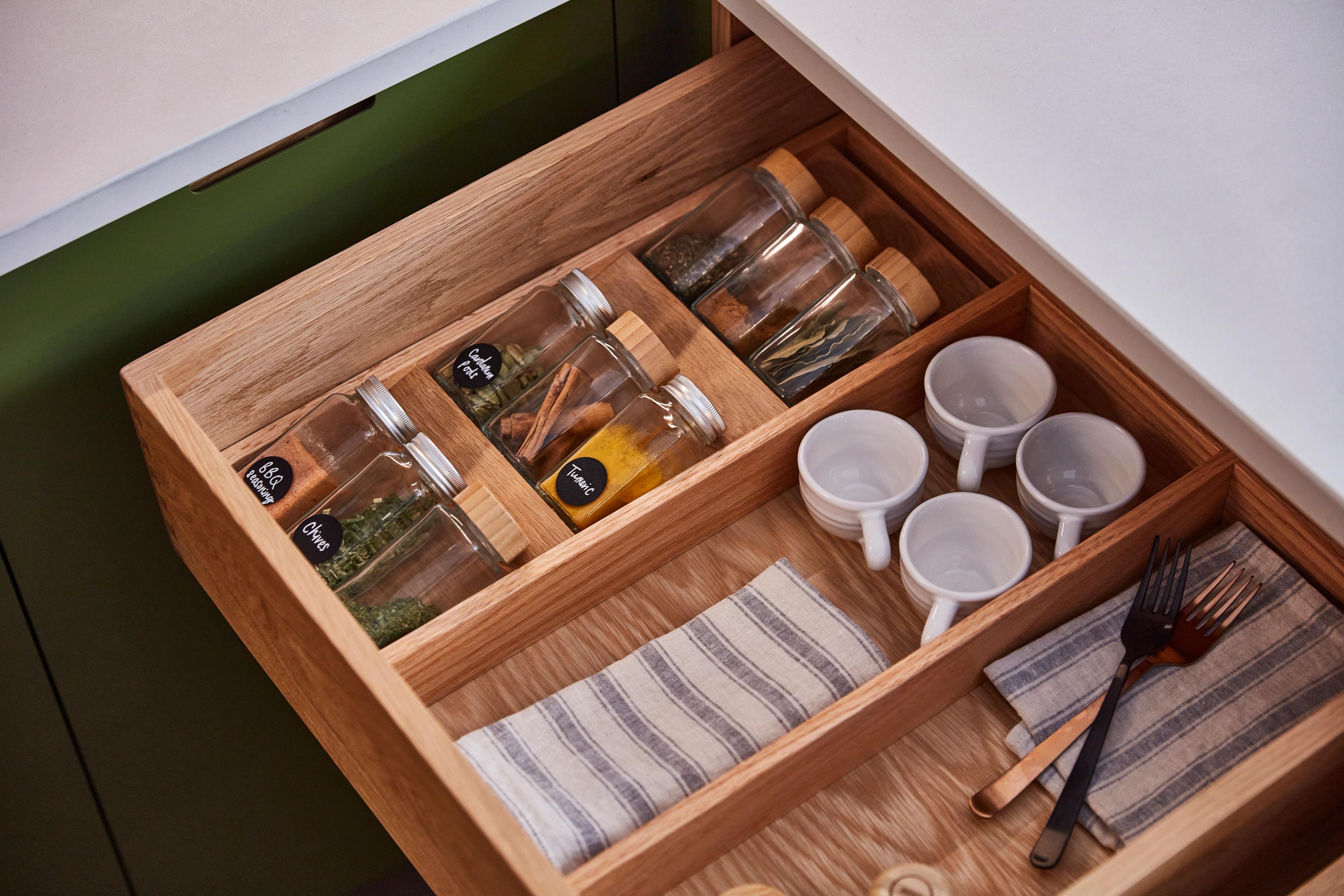 Open kitchen drawer with wooden dividers, holding glass jars with spices and four white mugs. A striped cloth and utensils like forks and a knife are neatly placed alongside. The drawer is under a green counter.