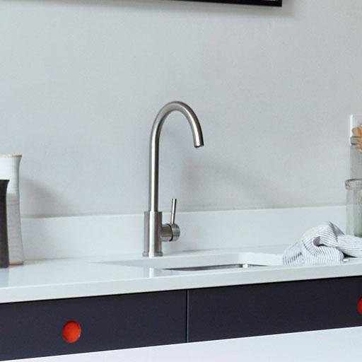 A modern kitchen countertop with a sleek, stainless steel faucet, a striped dish towel, a glass jar, a utensil holder, and white cabinetry with dark drawers featuring round orange handles.