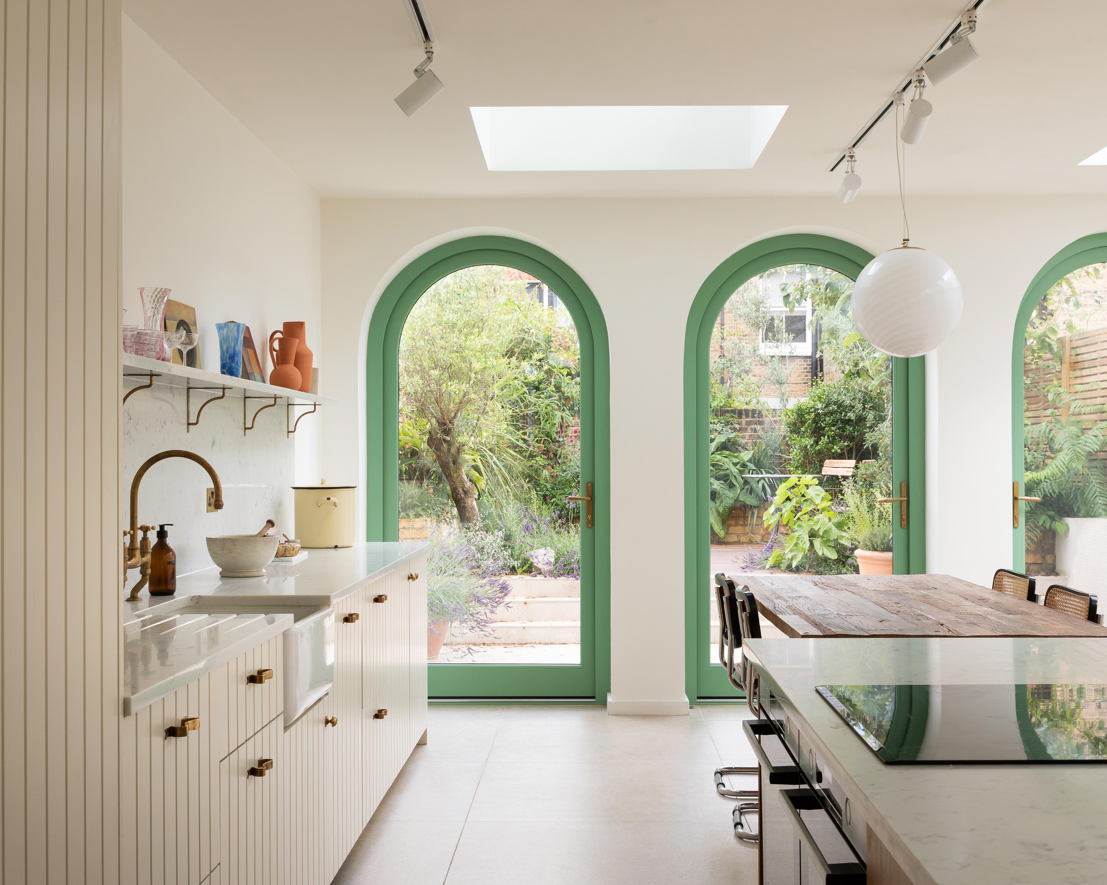 Bright kitchen with white cabinets and a large wooden table. Three arched green-framed glass doors lead to a lush garden. A round pendant light hangs from the ceiling, and a skylight adds natural light. Track lighting is installed above.