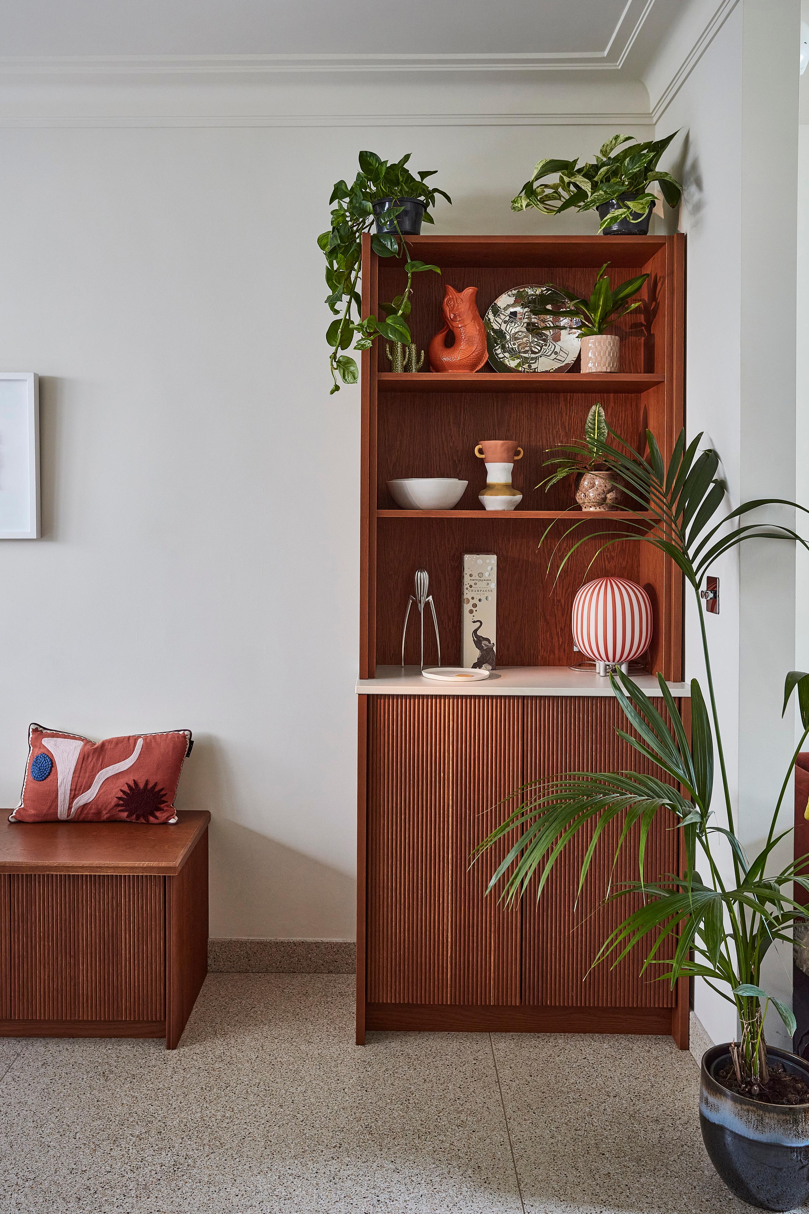 A wooden cabinet with plants, ceramic vases, and decor on its shelves stands by a white wall. A large potted plant is in front, and a bench with a patterned pillow is on the left. The scene is minimal and modern.