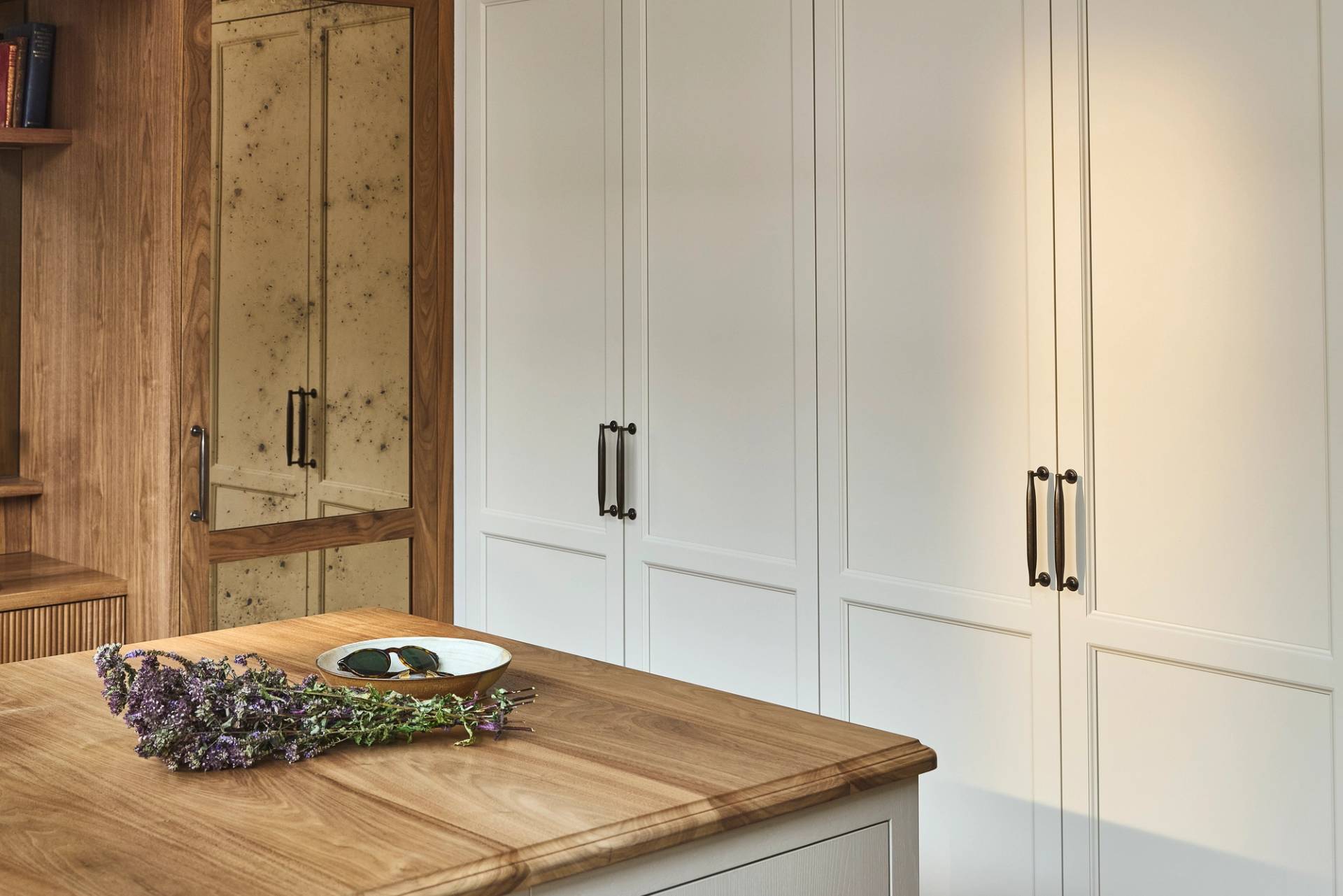 A modern kitchen with white paneled cabinets, black handles, and wood accents. A kitchen island with a wood countertop holds a plate with two dark stones and a bundle of purple flowers.