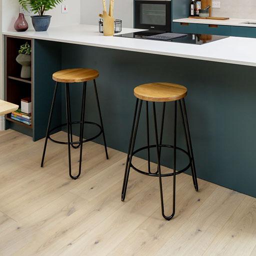 Two modern bar stools with round wooden seats and black metal legs are placed in front of a kitchen island with a white countertop. The kitchen features light wood flooring and minimalistic decor.