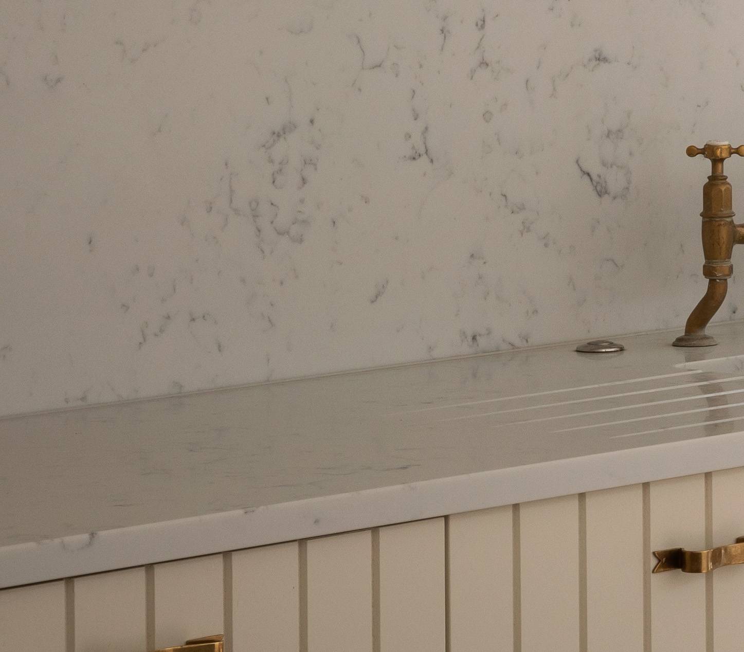 Close-up of a light-colored sink with a grooved draining board, marble countertop and backsplash, and a vintage-style brass faucet. The lower cabinet has vertical paneling and a gold handle.