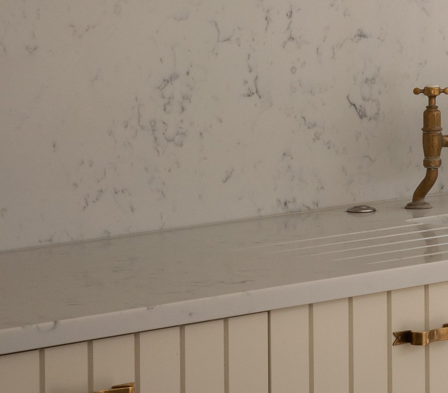 Close-up of a light-colored sink with a grooved draining board, marble countertop and backsplash, and a vintage-style brass faucet. The lower cabinet has vertical paneling and a gold handle.