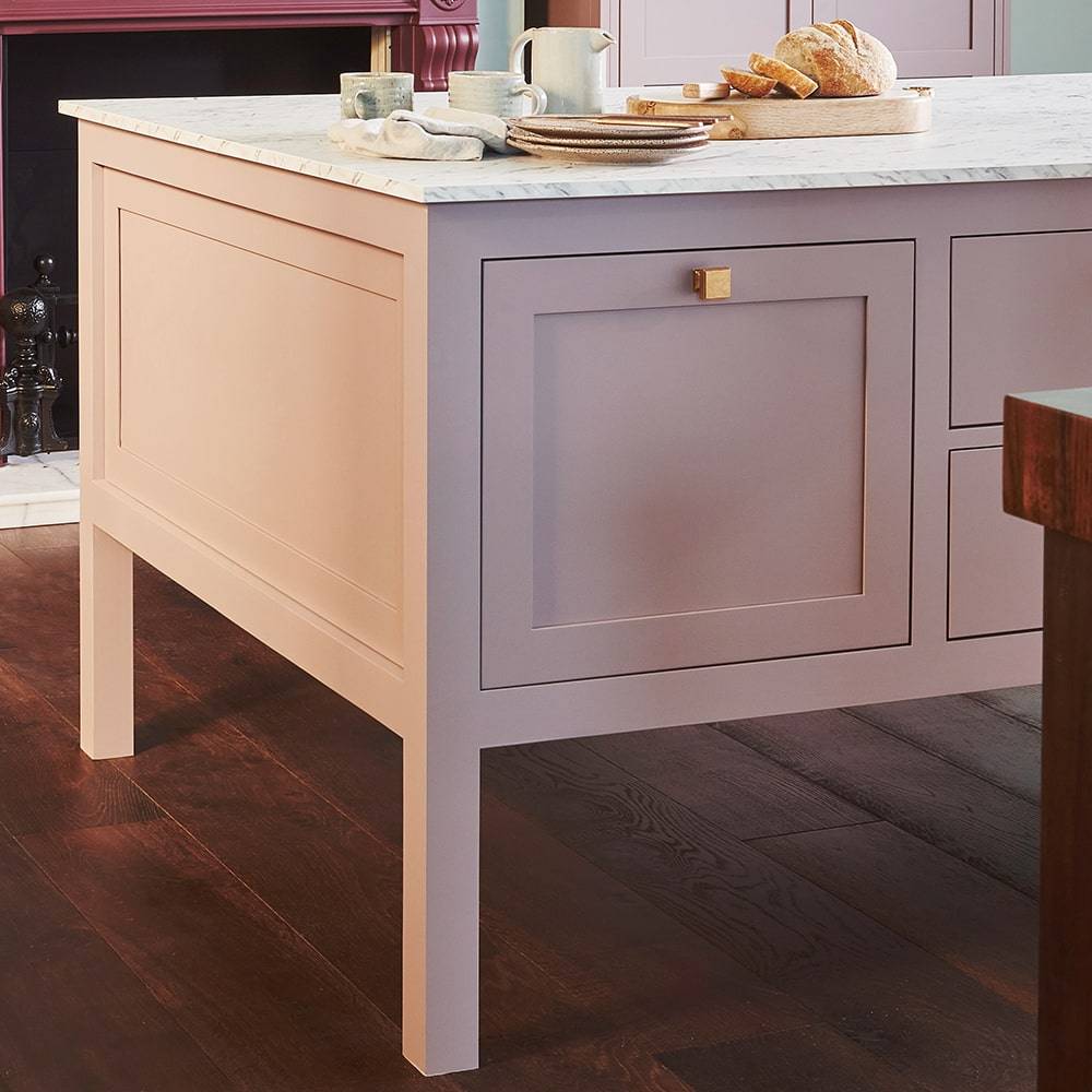 A light pink kitchen island with a marble countertop stands on a dark wooden floor. On the counter are a ceramic mug, a small plate, and a loaf of bread on a wooden board.