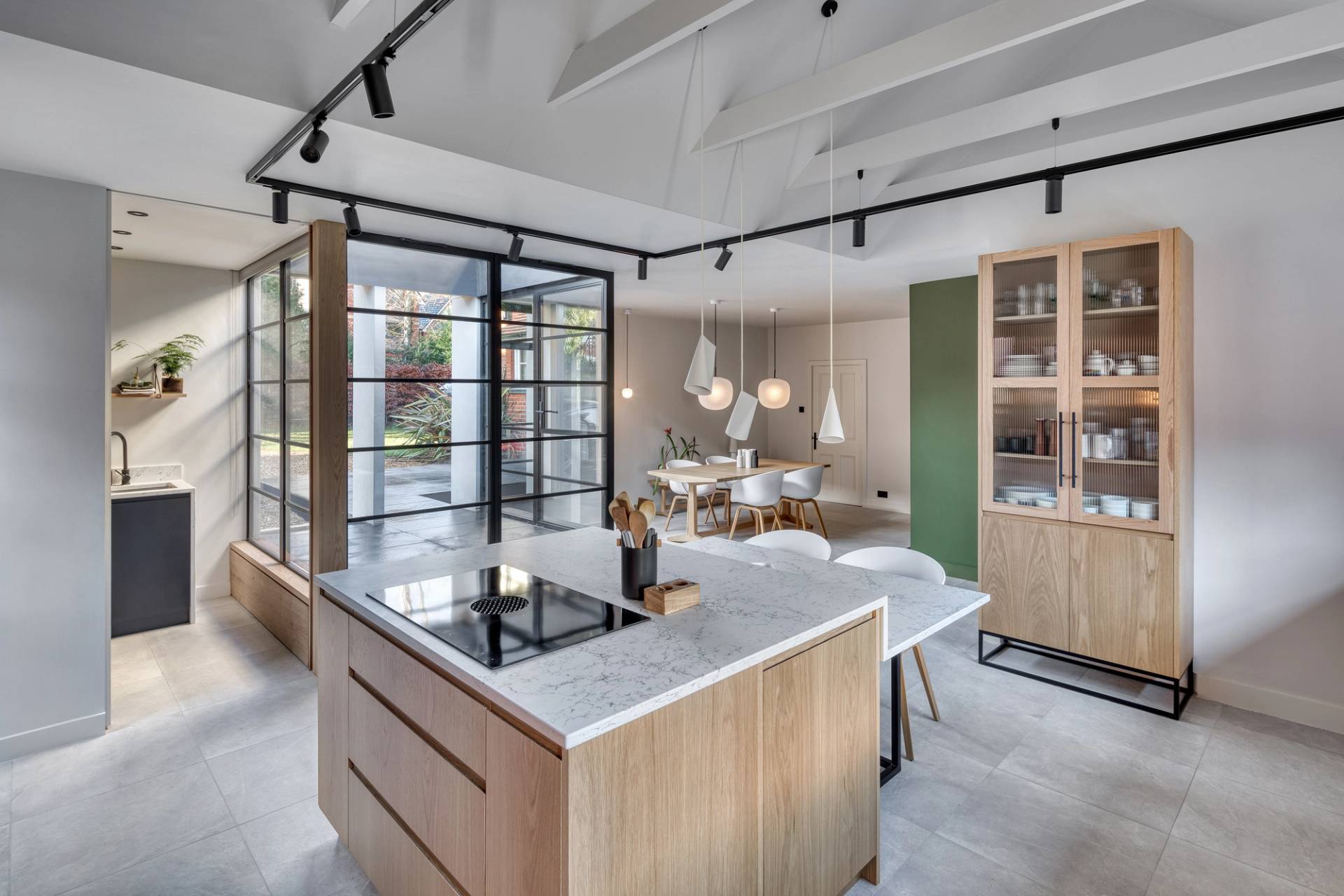 A modern kitchen and dining area with a marble island, wooden cabinets, and a built-in stovetop. The open layout features large windows, a green accent wall, and minimalist pendant lights over a dining table with white chairs